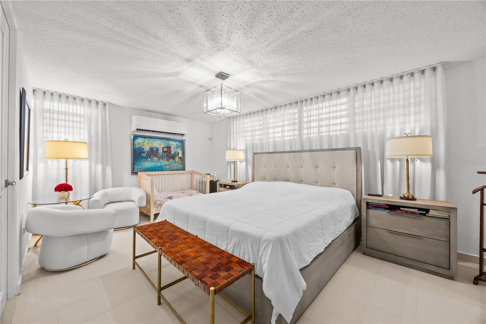 Furnished bedroom with an upholstered bed, sheer curtains, a crib, and a woven bench.