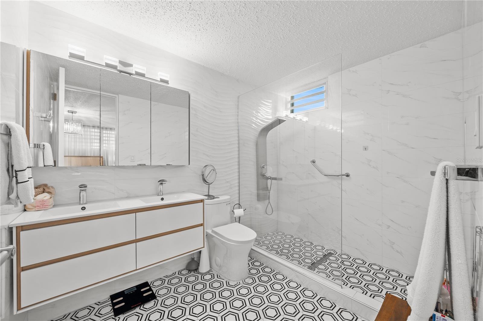 Modern bathroom with double vanity, glass walk-in shower, marble-look walls, and geometric tile floor.