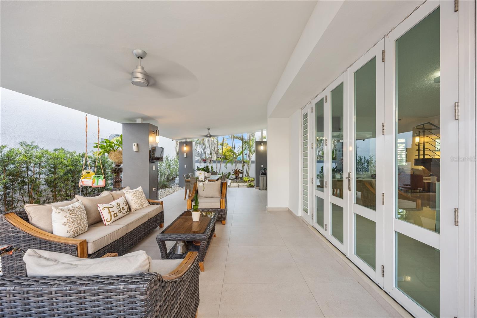 Covered patio with wicker seating, tiled floor, and glass doors beside a tropical garden.