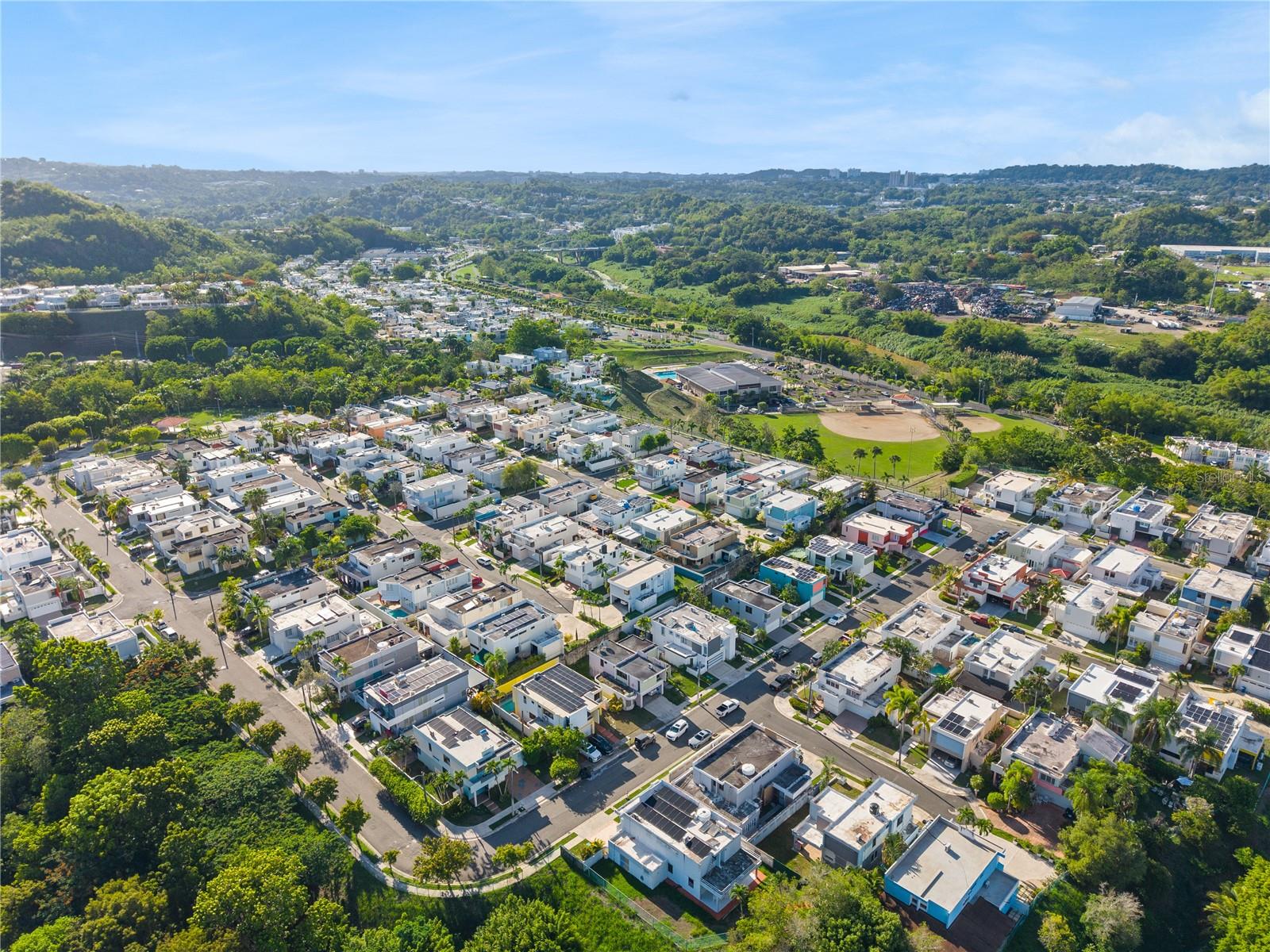 Aerial view of a residential neighborhood bordered by wooded hills, with a baseball field in the distance.