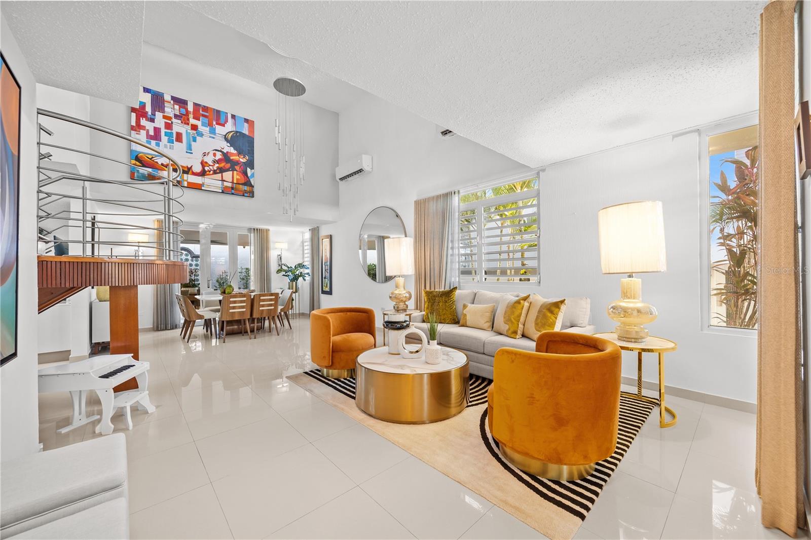 Open-plan living and dining room with orange chairs, a gray sofa, glossy tile floors, and a curved staircase.