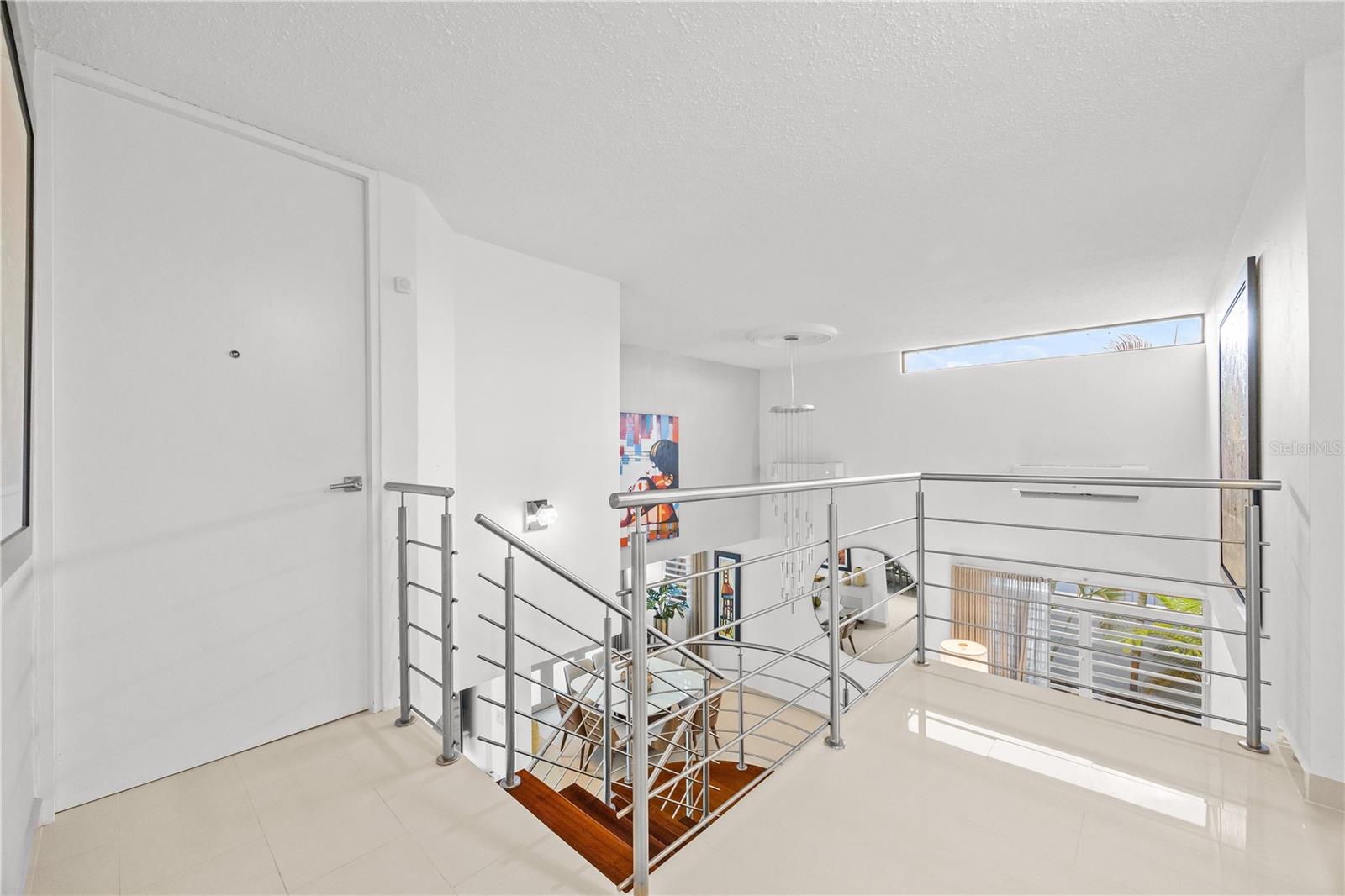 Bright upstairs landing overlooking a double-height living area, with a stainless-steel stair railing and white tile floors.