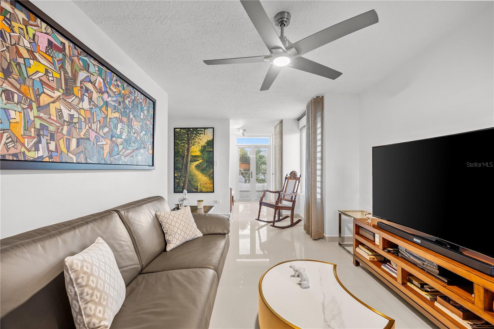 Living room with a leather sofa, colorful wall art, rocking chair, and large television.