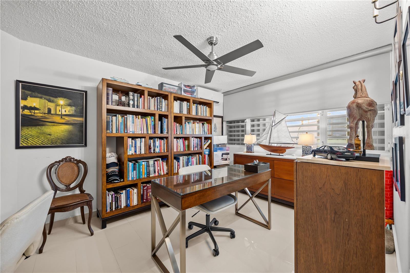 Home office with book-filled wooden shelves, a glass-top desk, and louvered windows.