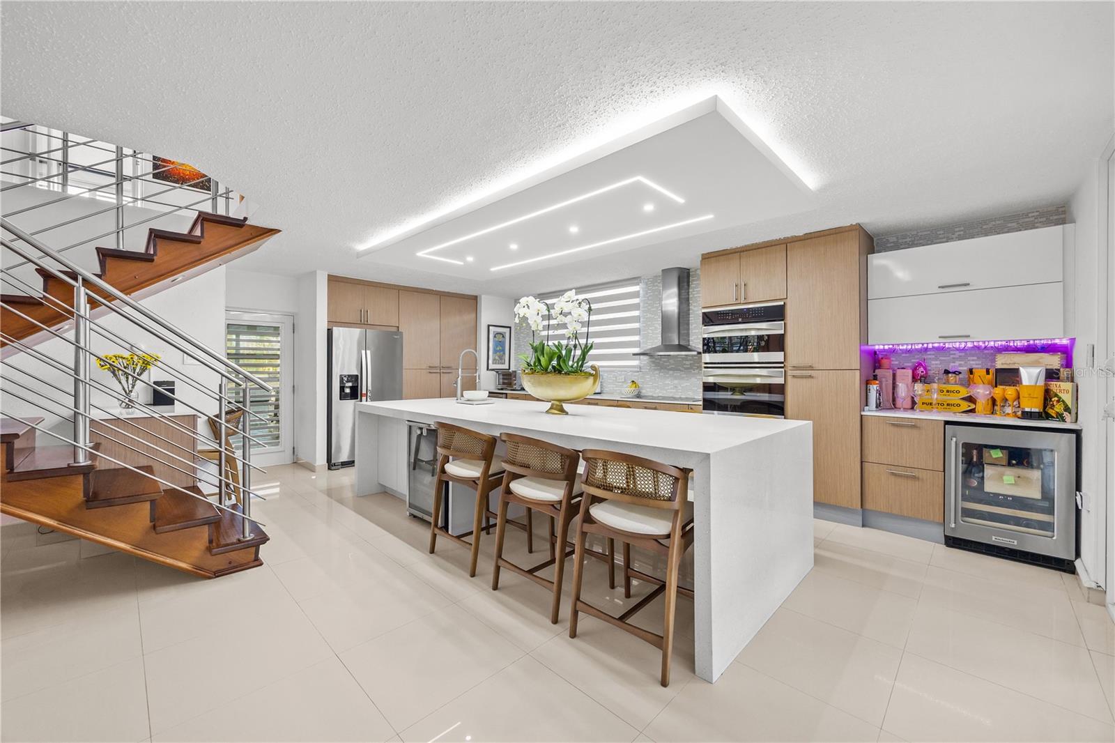 Modern kitchen with a large white island, wood cabinetry, stainless appliances, and a sculptural staircase.
