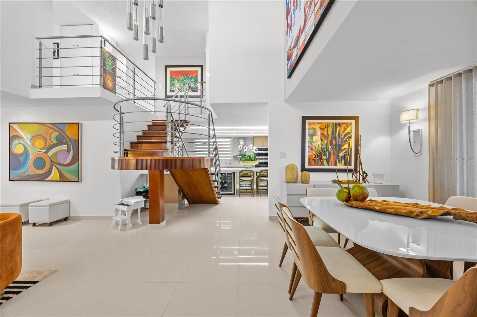 Open-plan dining room with a curved wood staircase, colorful artwork, and kitchen beyond.