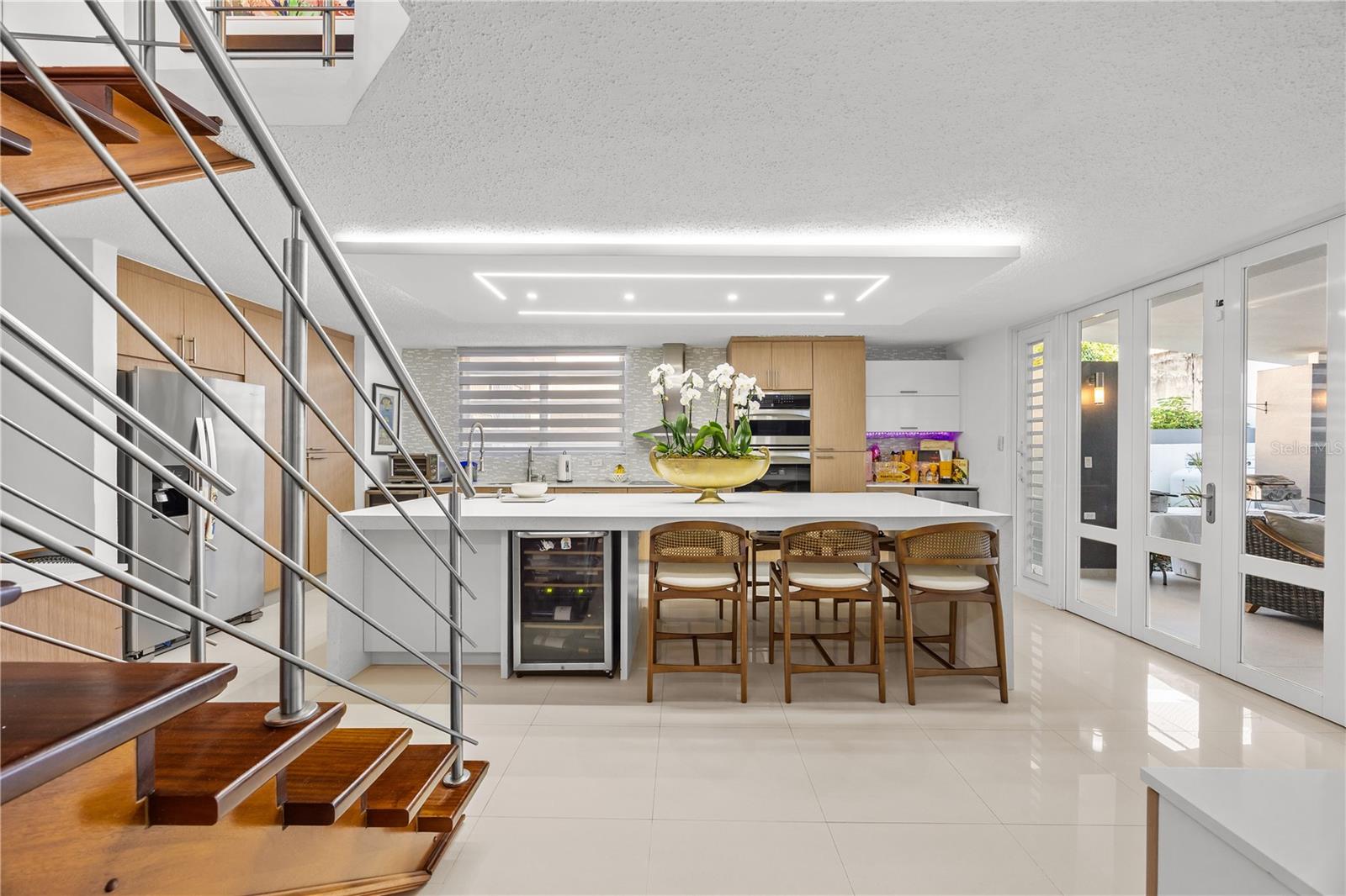 Modern kitchen with a white island and bar stools beside a wood staircase and glass patio doors.