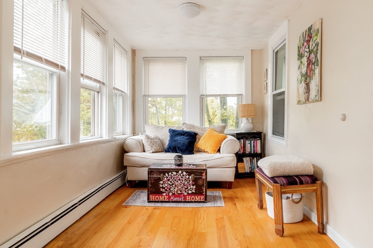 Sunroom with a cream sofa, colorful pillows, hardwood floors, and windows overlooking trees.