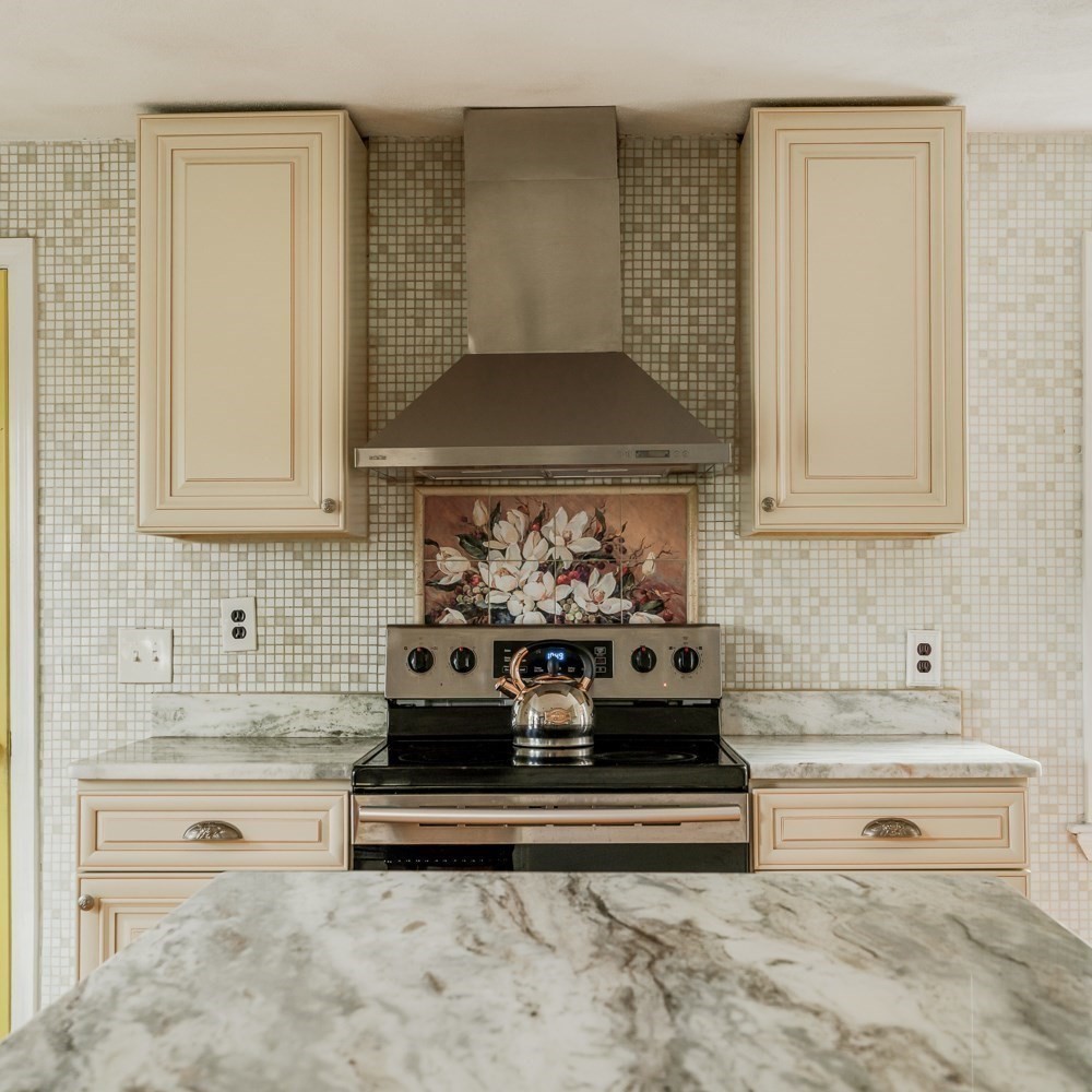 Kitchen with cream cabinets, a stainless steel range, floral backsplash accent, and marble countertops.