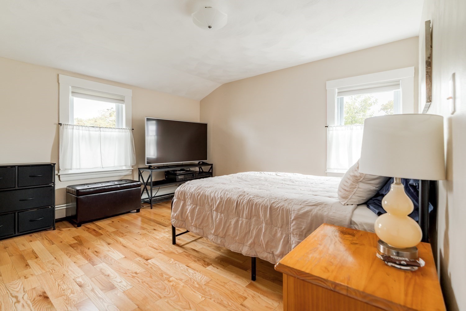 Furnished bedroom with a large bed, hardwood floors, and two curtained windows.