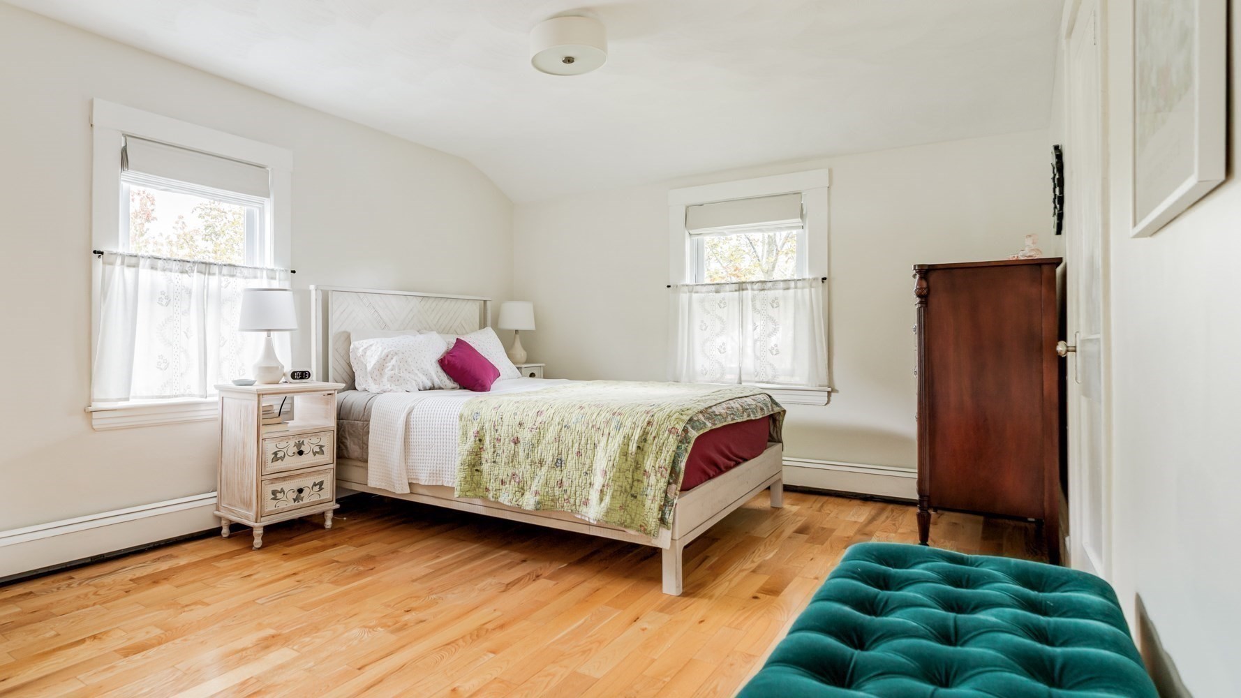 Furnished bedroom with hardwood floors, floral bedding, two bright windows, and a teal tufted bench.