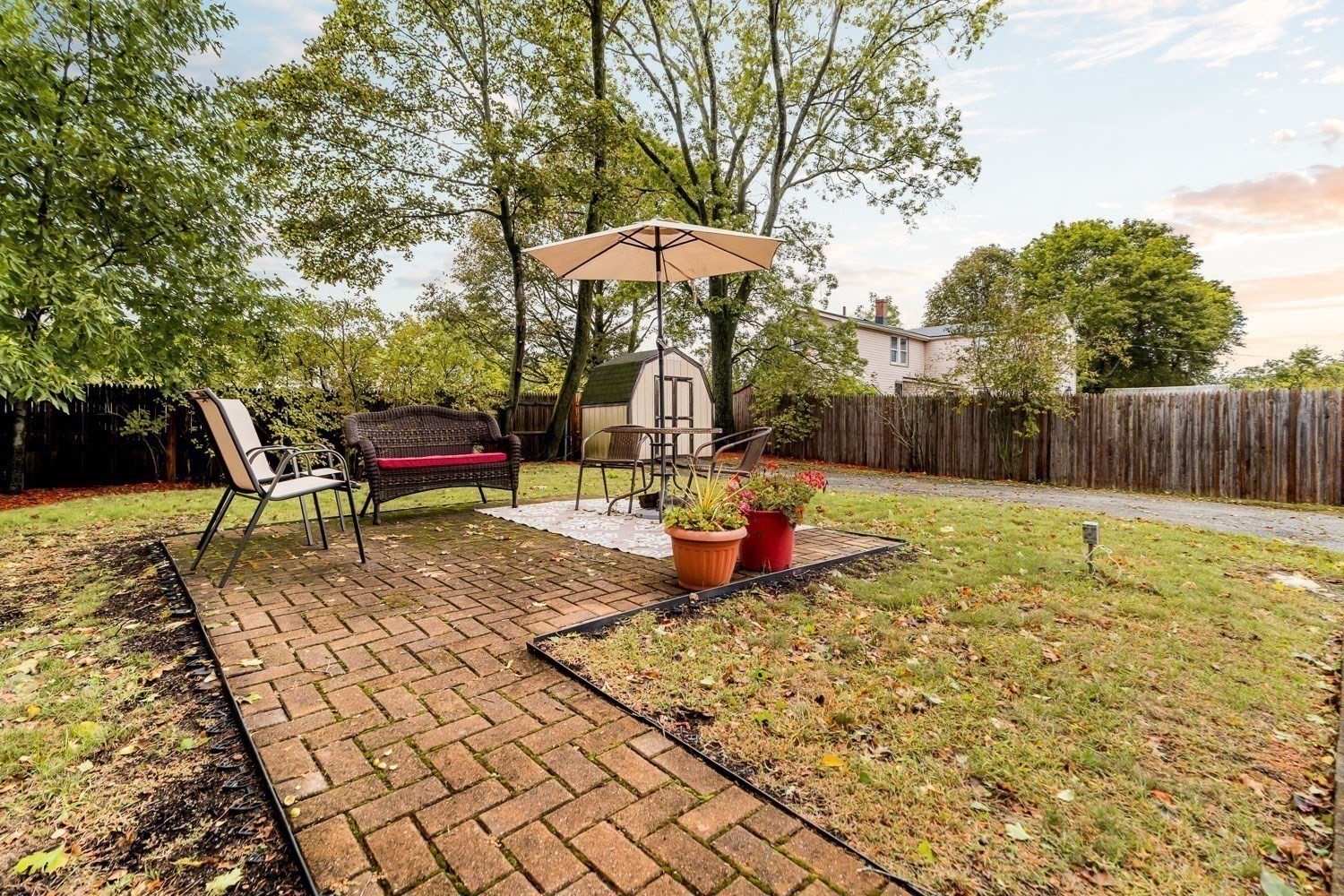 Brick backyard patio with outdoor seating beneath an umbrella, potted plants, and a wooden fence.
