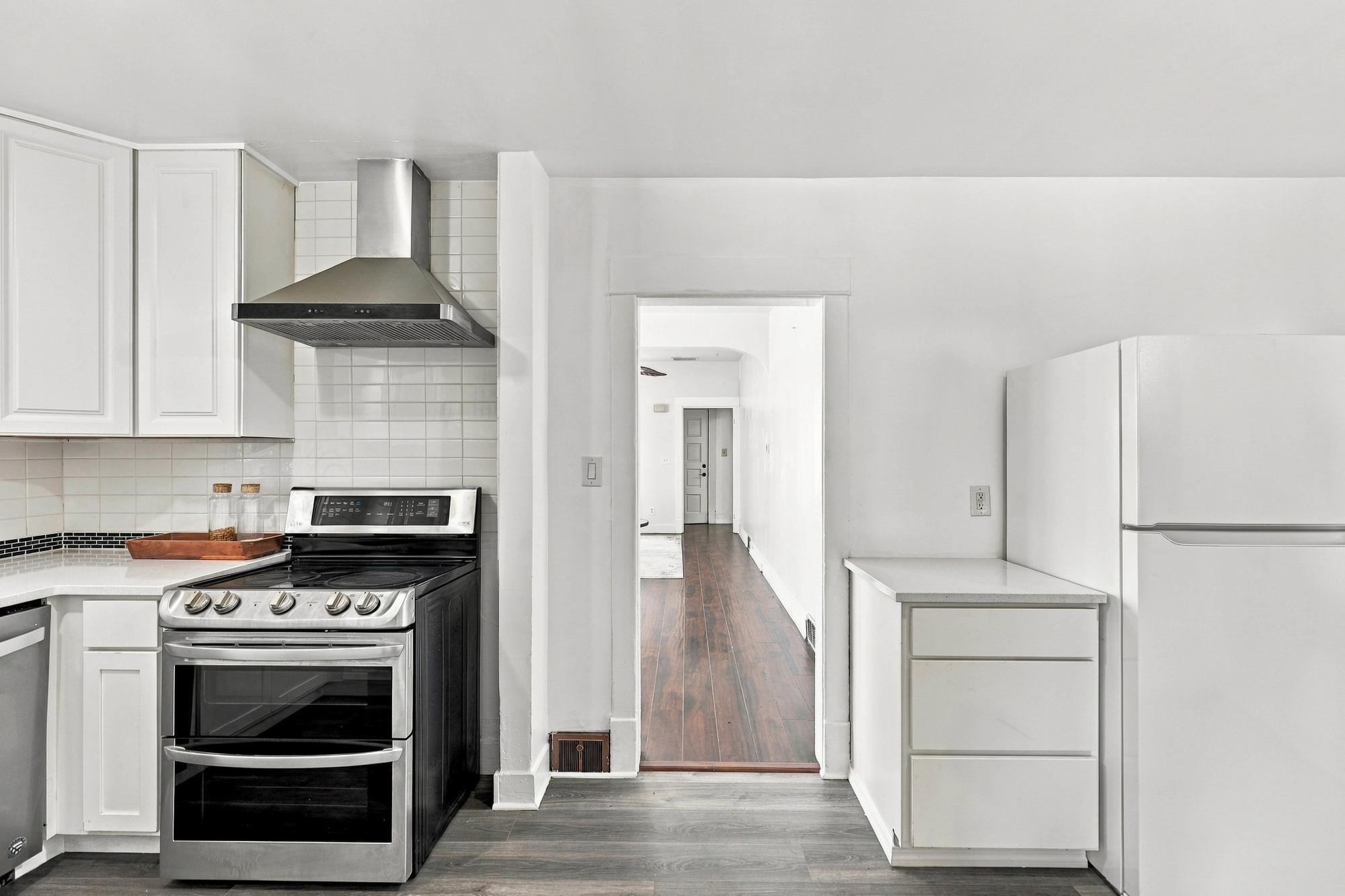 White kitchen with stainless range and tiled backsplash opening onto a long wood-floored hallway.
