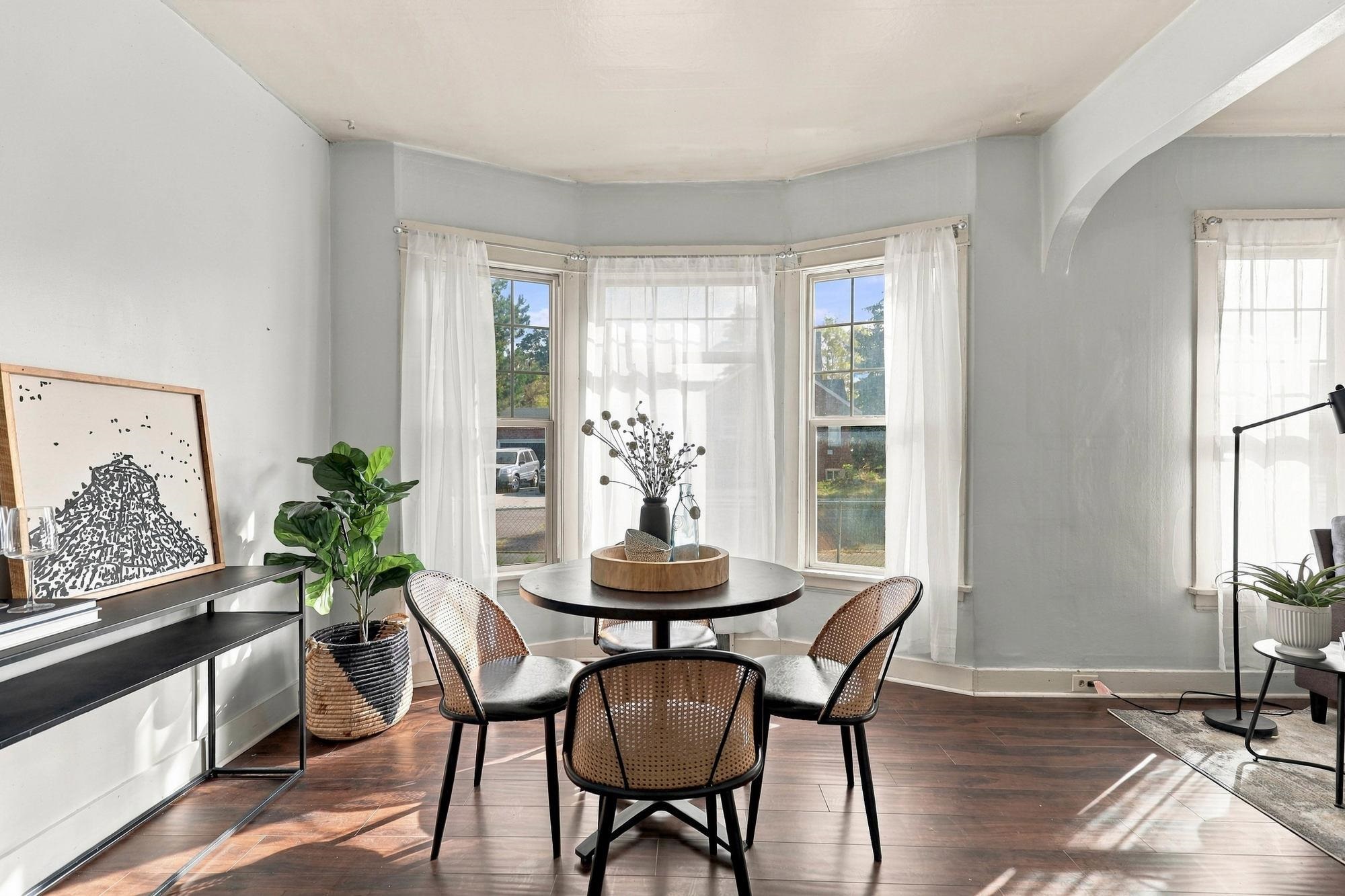 Dining area with a round table and woven chairs in front of a curtained bay window.