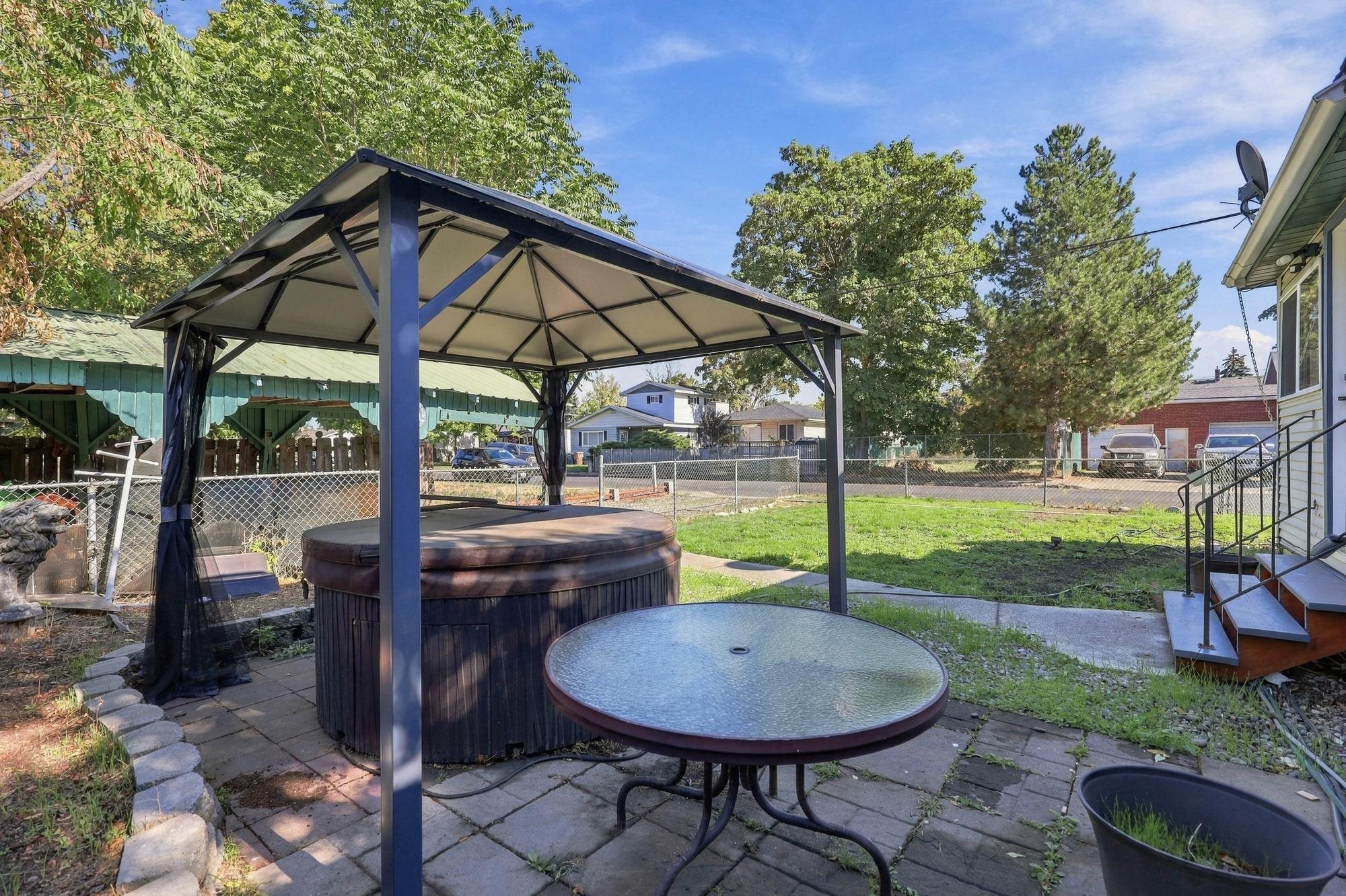 Backyard hot tub beneath a metal gazebo, with a round patio table and fenced lawn.