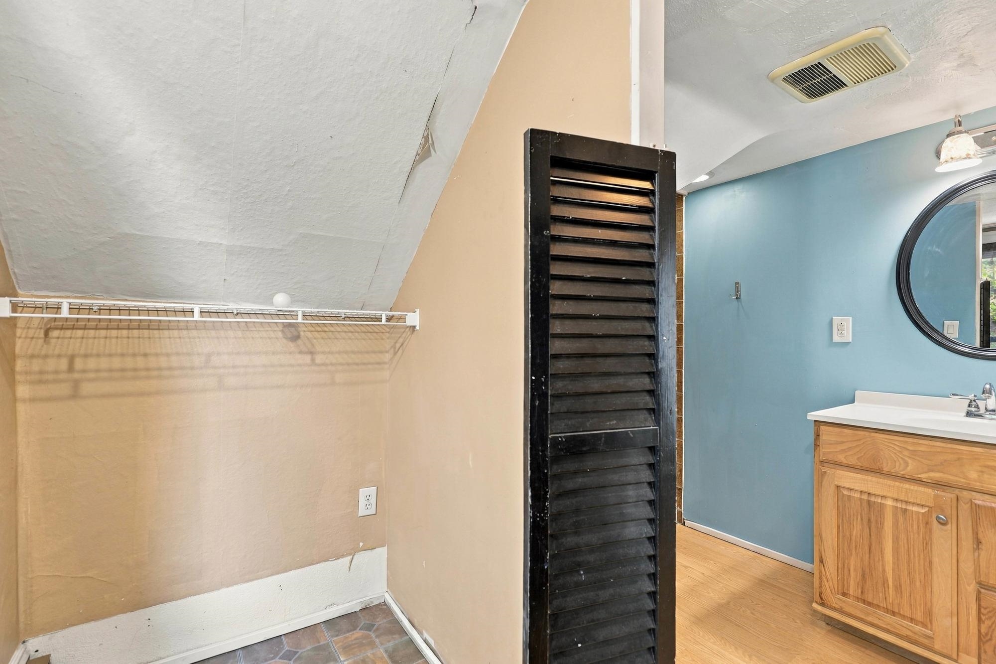 Bathroom with a wooden vanity, round mirror, and adjacent closet alcove with a black louvered door.