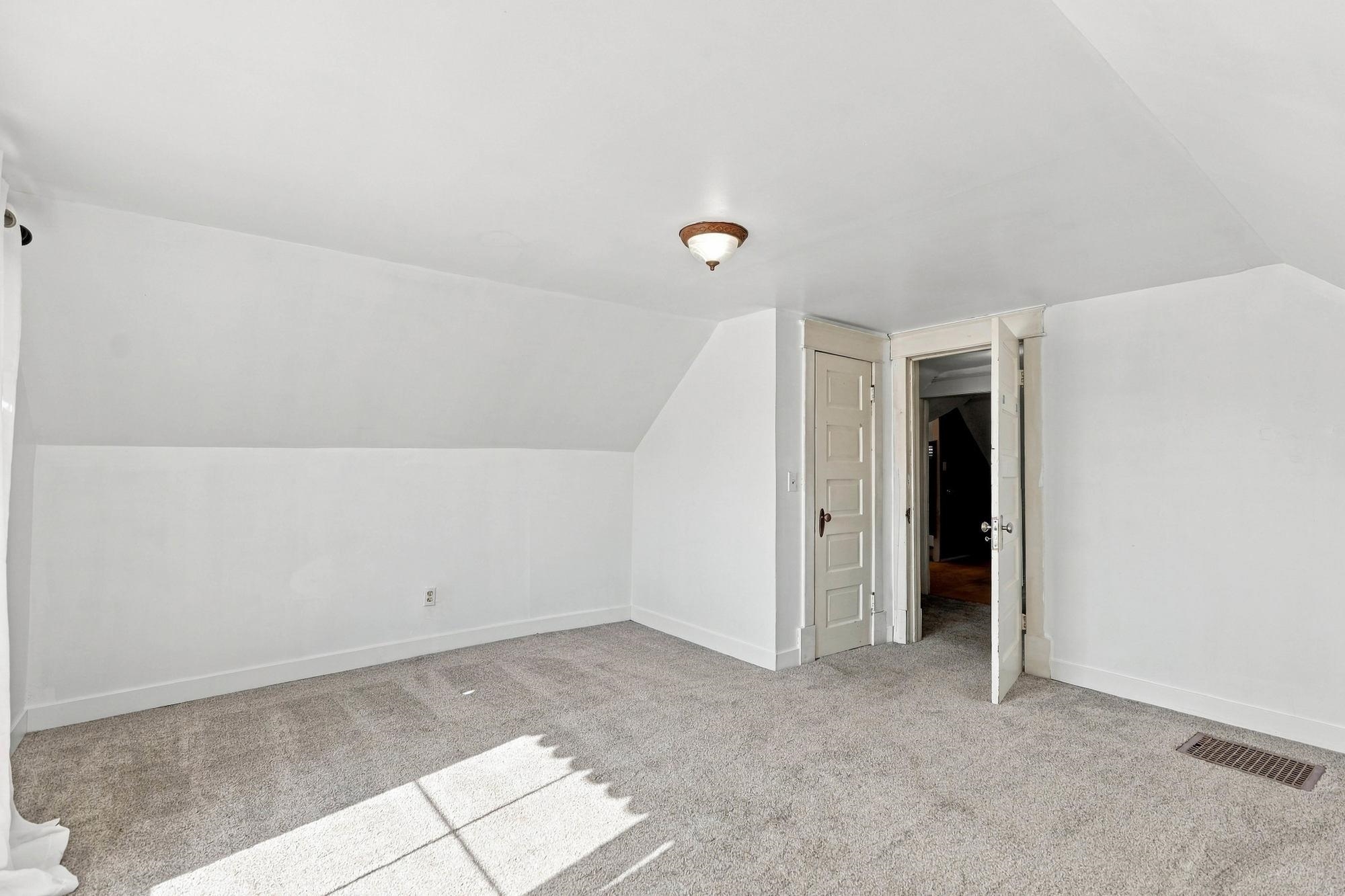 Empty carpeted room with a sloped ceiling, sunlit floor, and open doorway.