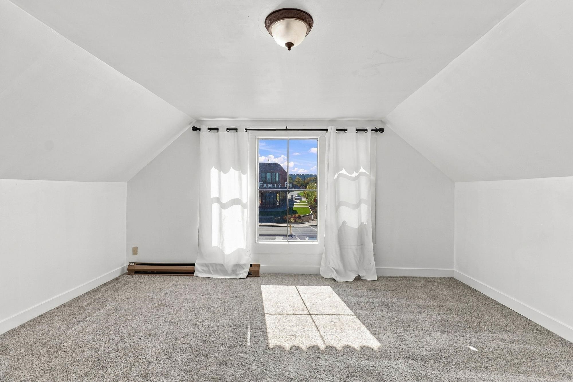 Empty carpeted room with sloped ceilings, white curtains, and sunlight streaming through the window.