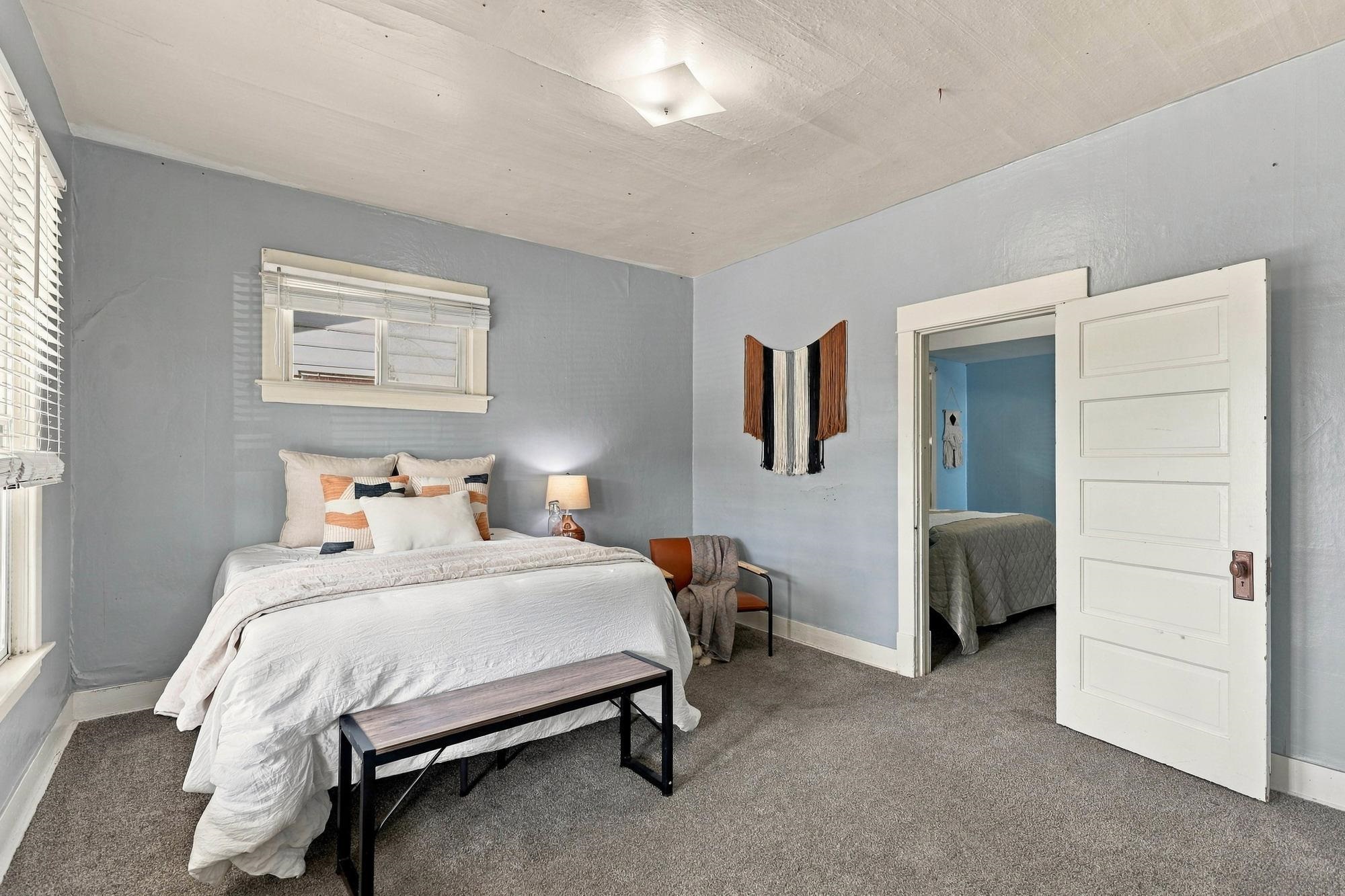 Staged bedroom with a white bed and bench; open doorway looks into a second bedroom.