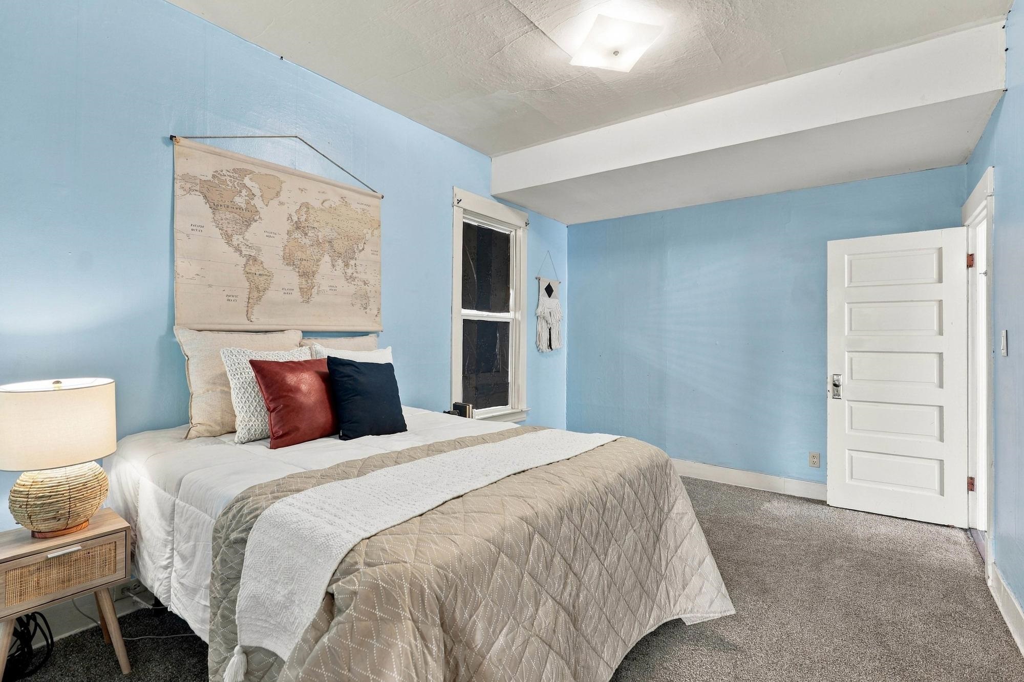 Staged bedroom with blue walls, a neatly made bed, world map wall hanging, and open white door.
