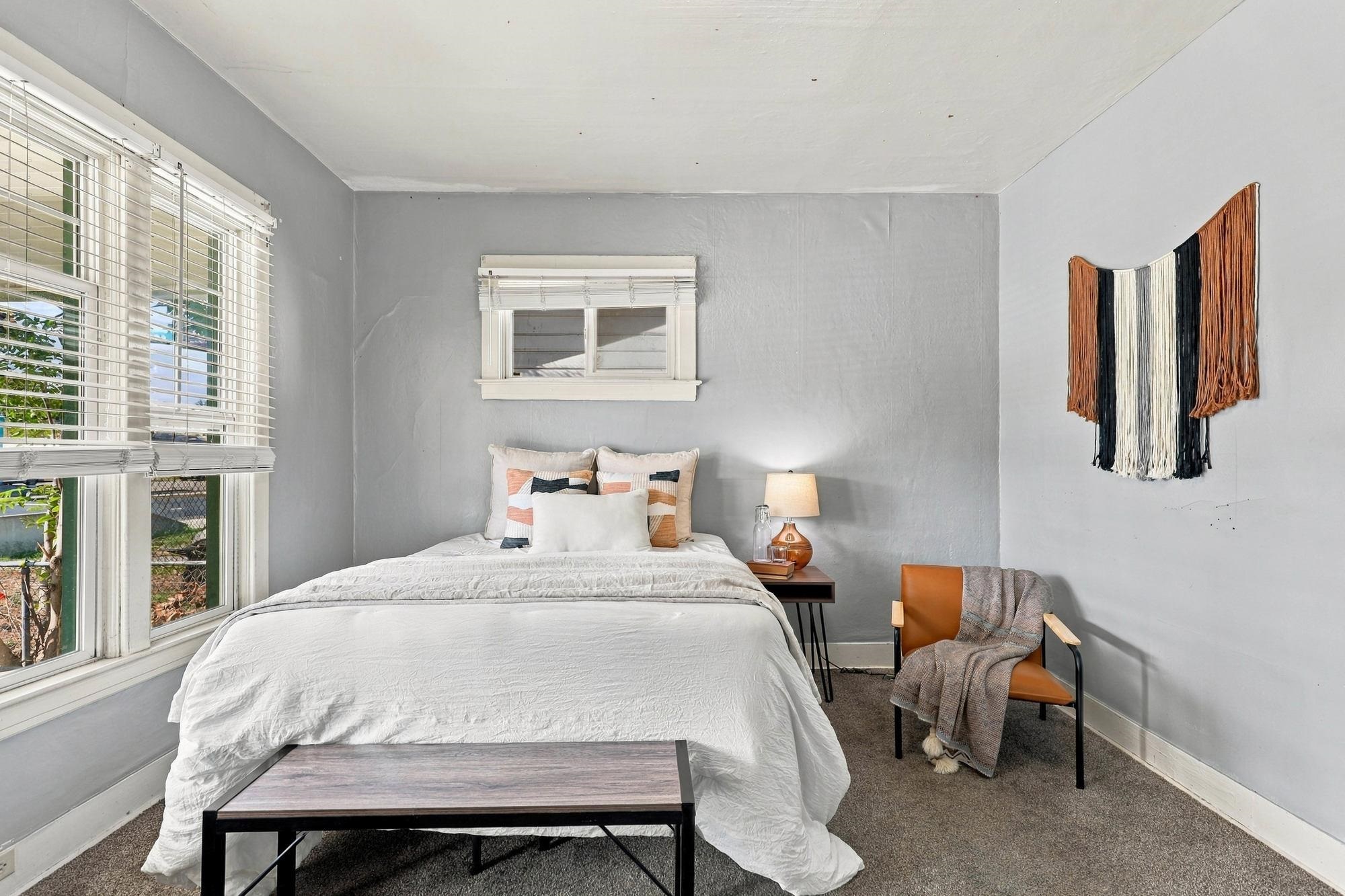 Furnished bedroom with a white bed, large windows with blinds, and a woven wall hanging.