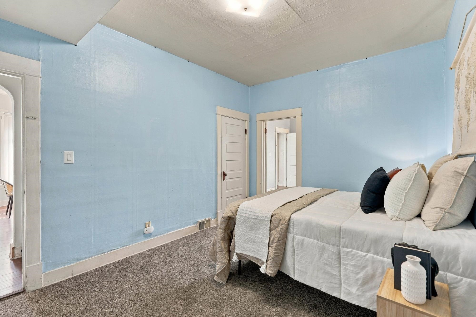 Staged bedroom with pale blue walls, a white bed, layered neutral bedding, and gray carpet.