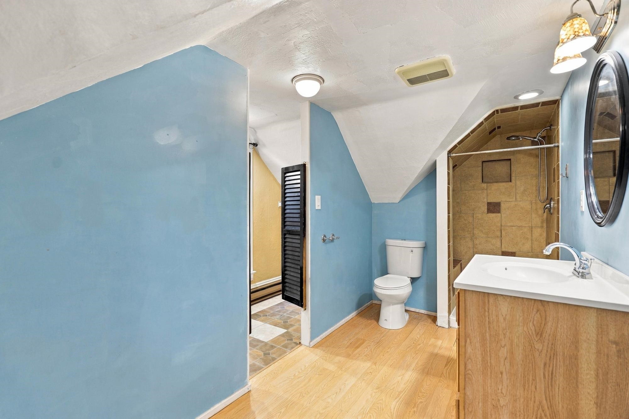 Bathroom with blue walls, a sloped ceiling, wood vanity, toilet, and tan tiled walk-in shower.
