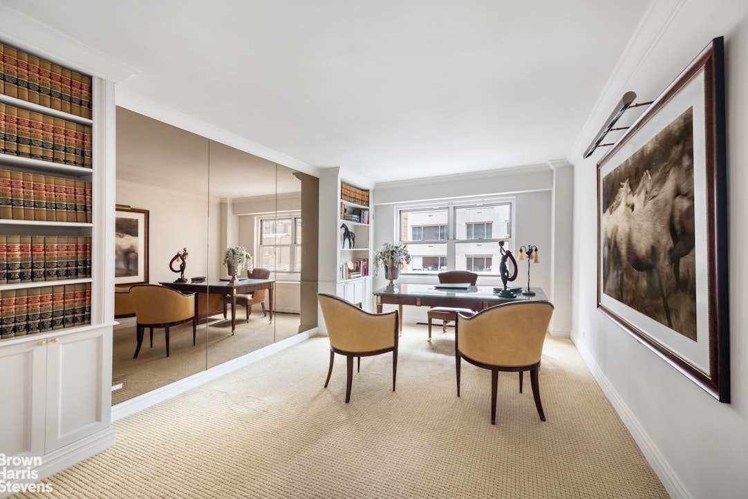 Home office with built-in bookshelves, mirrored wall, glass desk, and two upholstered chairs.