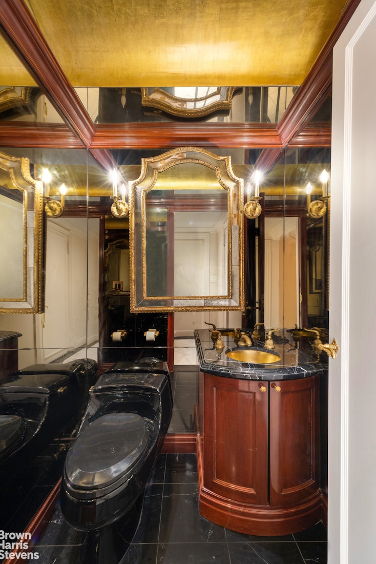 Bathroom with a curved dark wood vanity, black marble surfaces, mirrored walls, and an ornate gold-framed mirror.
