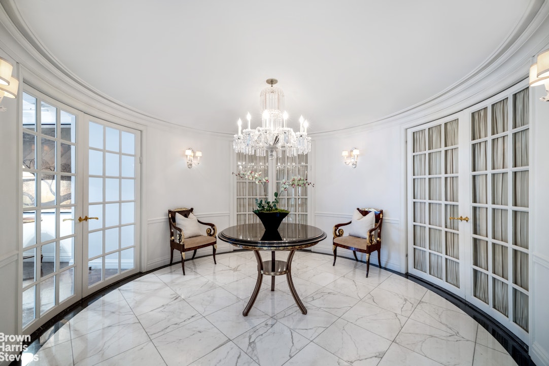 Curved dining room with a round table, crystal chandelier, French doors, and white marble flooring.