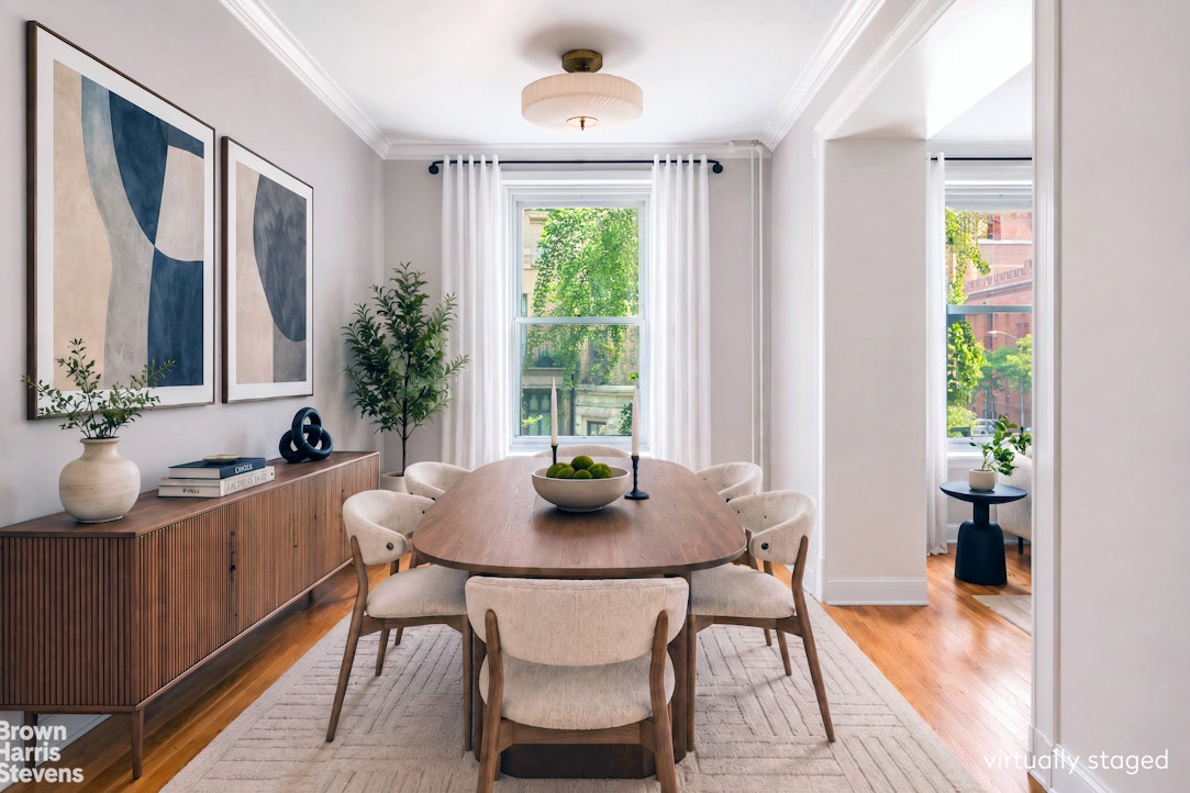 Dining room with an oval wood table, upholstered chairs, and tall windows overlooking greenery.