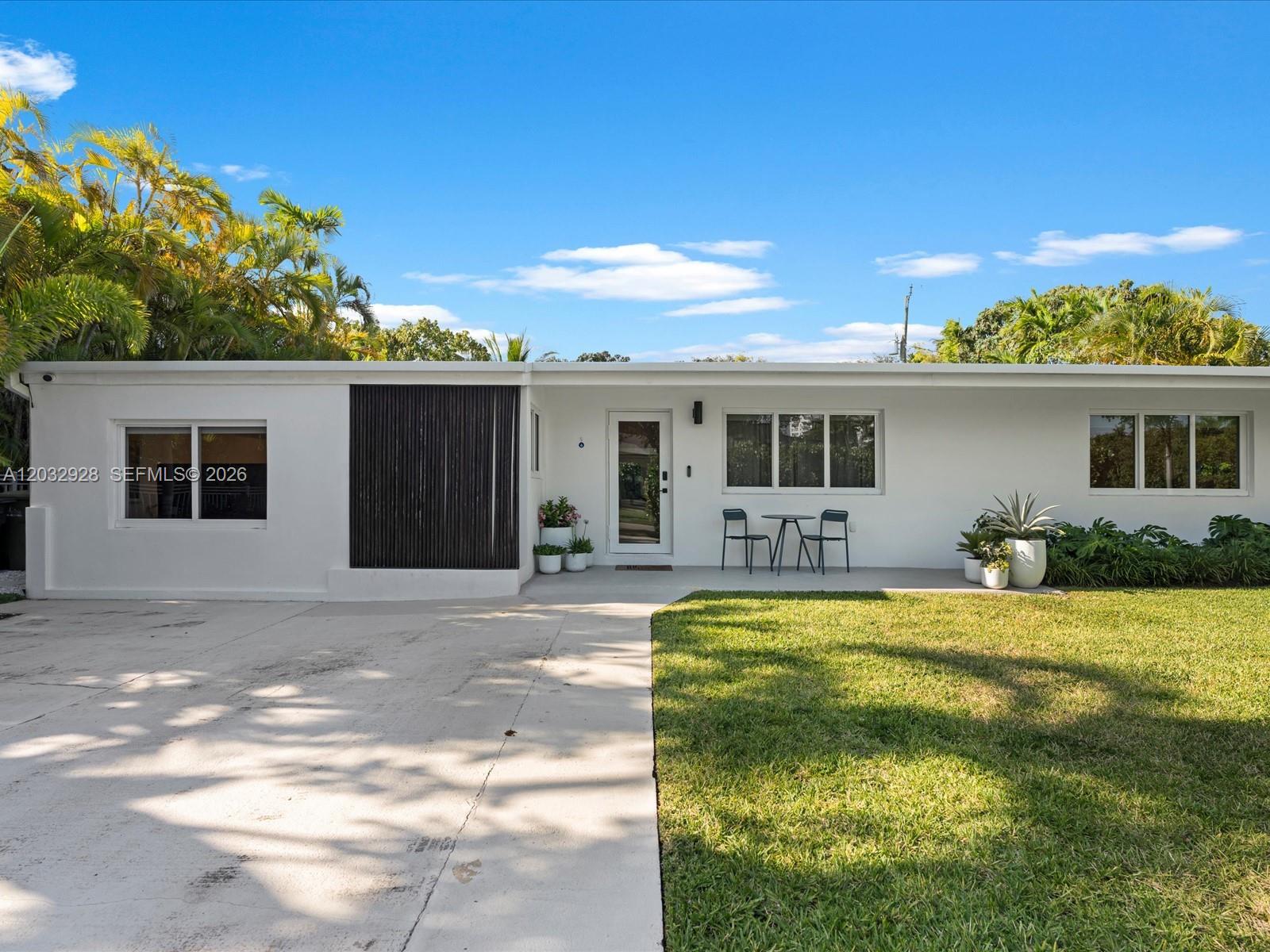 Modern single-story white house with a flat roof, dark facade panel, driveway, lawn, and tropical plants.