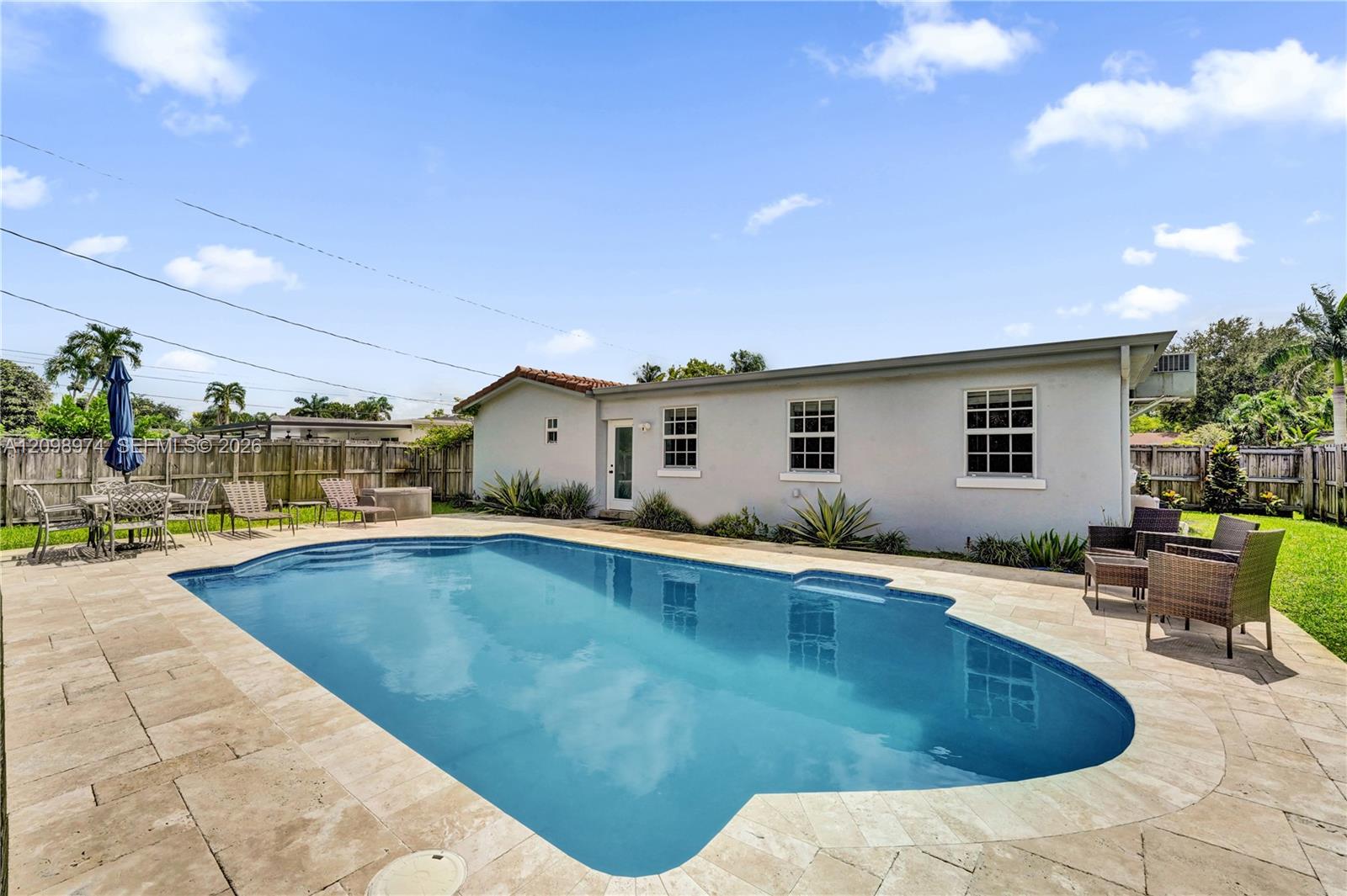 Backyard pool beside a paved patio with outdoor seating and a fenced single-story home.