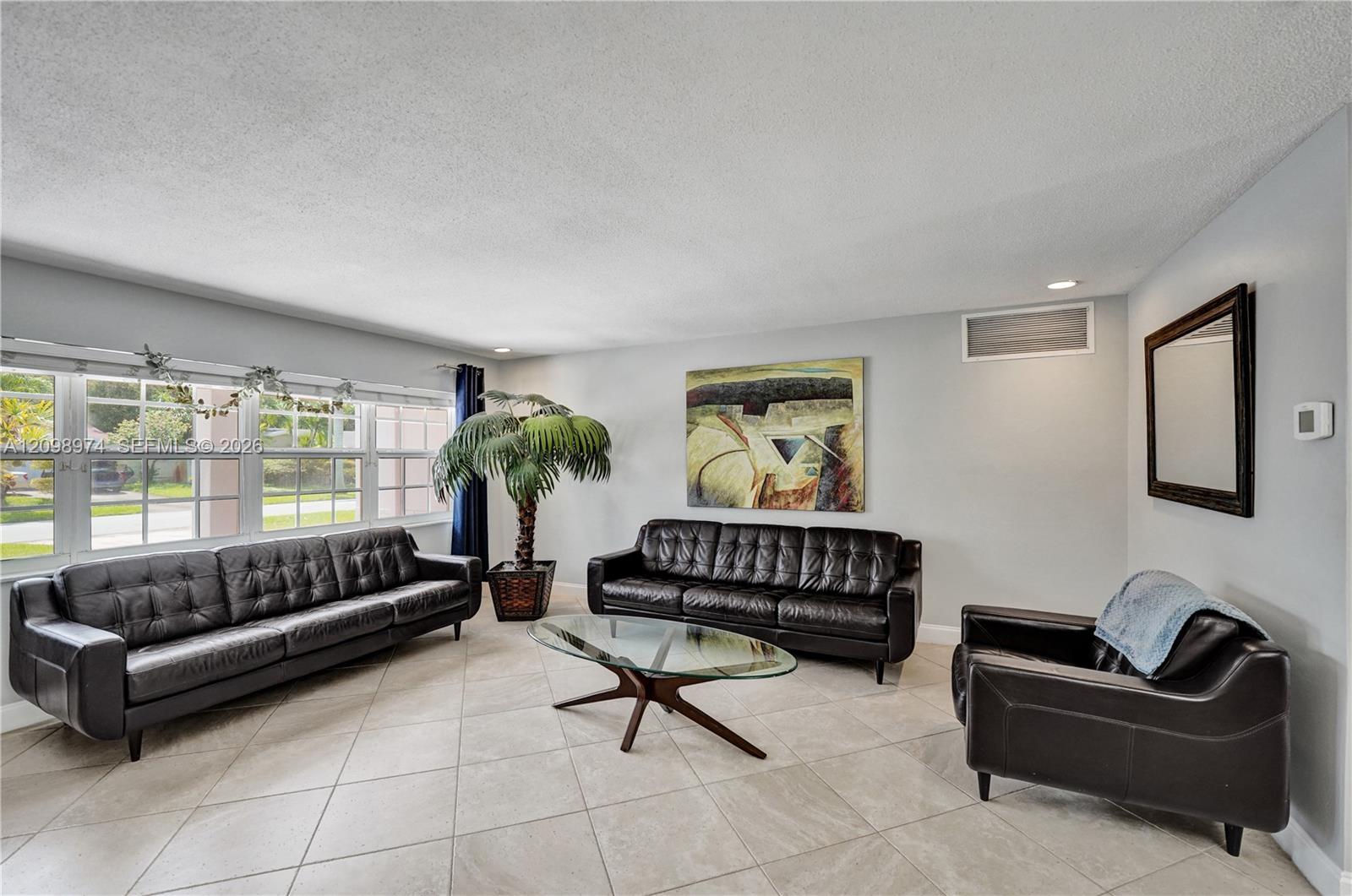 Living room with black leather seating, a glass coffee table, and large windows overlooking a yard.