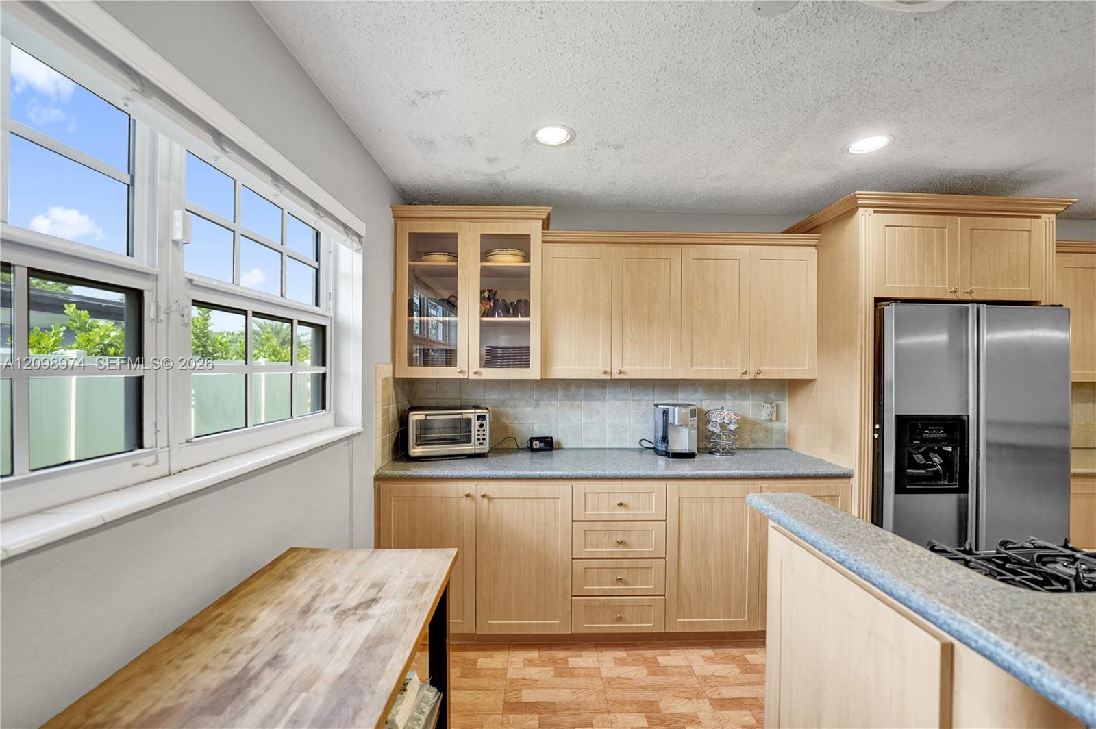 Kitchen with light wood cabinetry, stainless steel refrigerator, gas cooktop, and large windows.