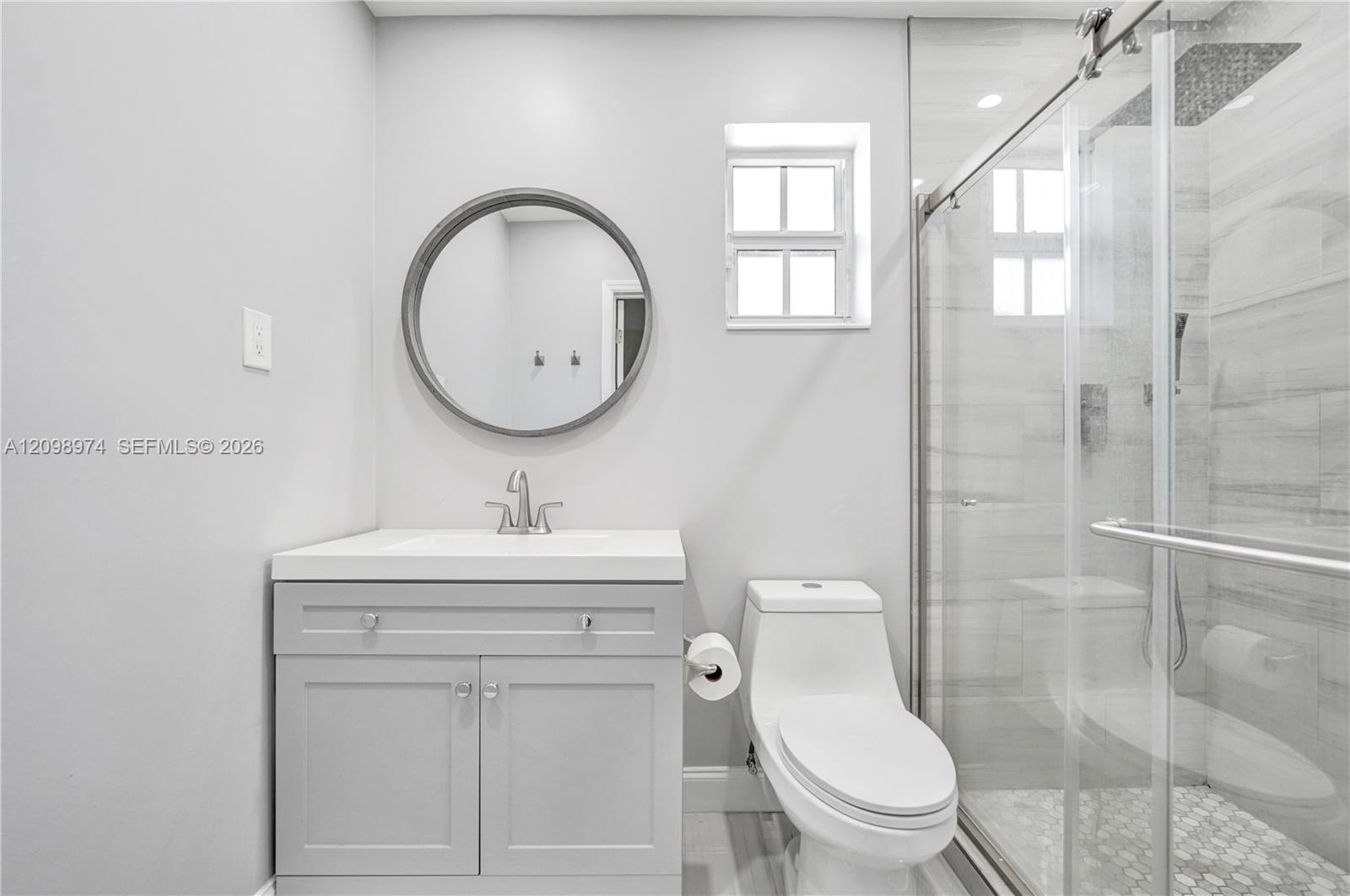 Bathroom with a white vanity, round mirror, toilet, and glass-enclosed tiled shower.