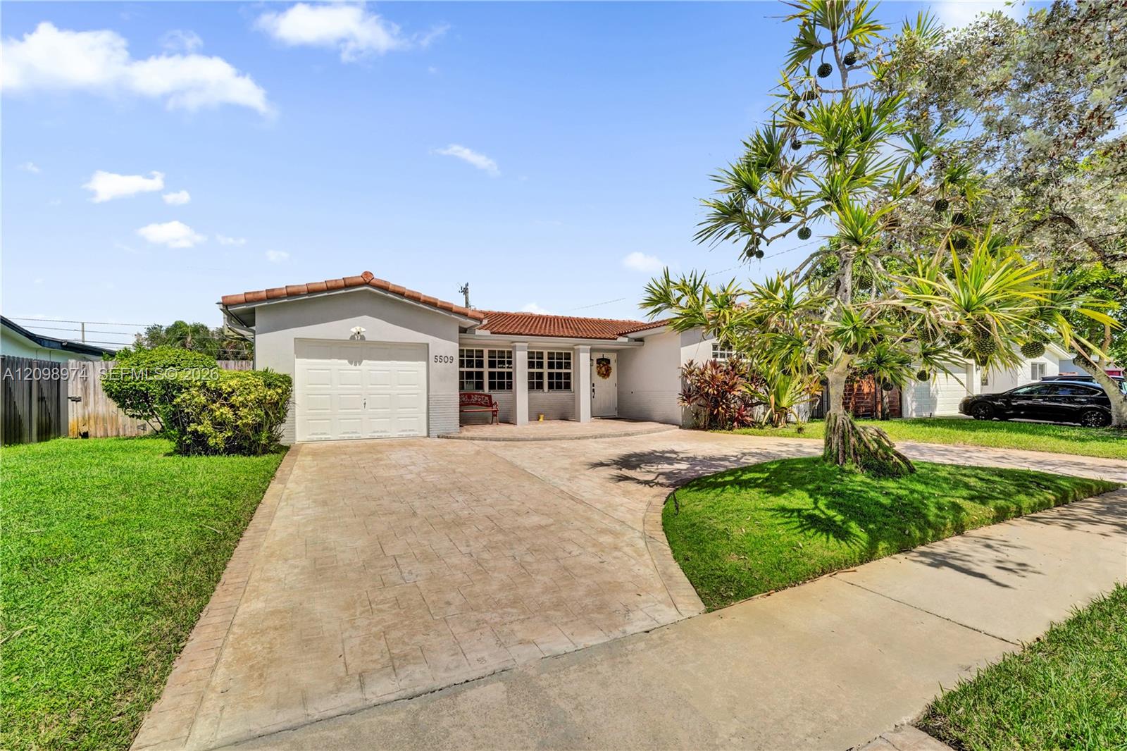 Single-story home with a red tile roof, broad driveway, attached garage, and tropical landscaping.