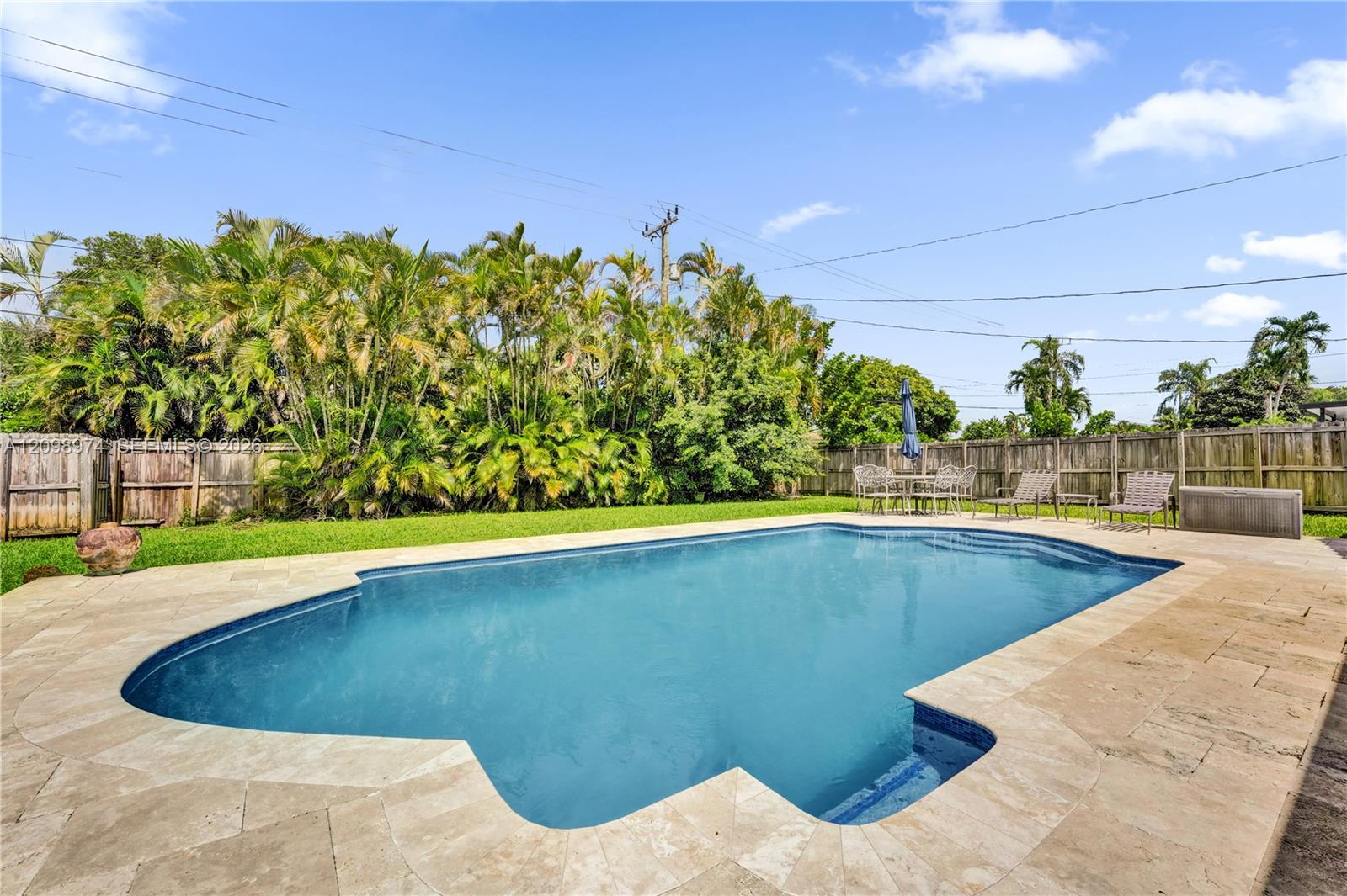 Backyard swimming pool with a pale stone patio, tropical landscaping, and outdoor seating.