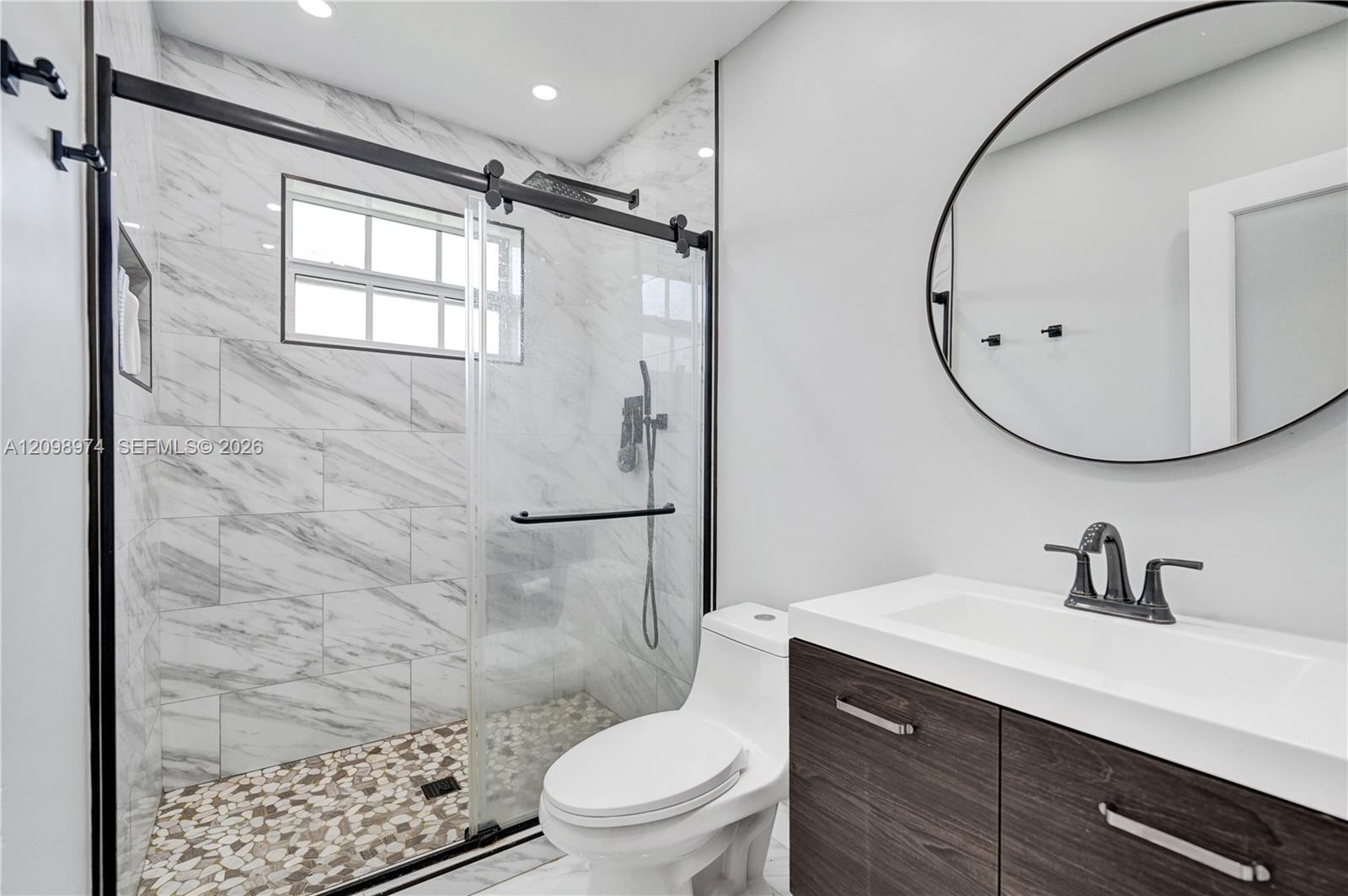 Modern bathroom with a glass shower, pebble-mosaic floor, dark wood vanity, and round mirror.