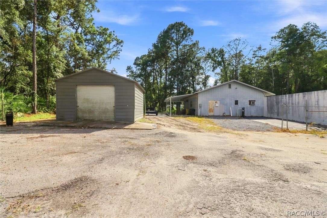 Detached gray garage and large metal outbuilding beside a gravel lot surrounded by trees.