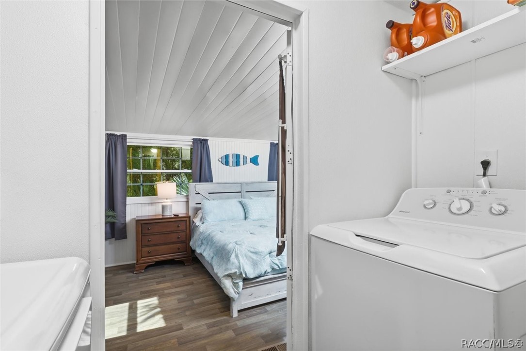 Laundry area opens through a doorway to a furnished bedroom with a blue bedspread.