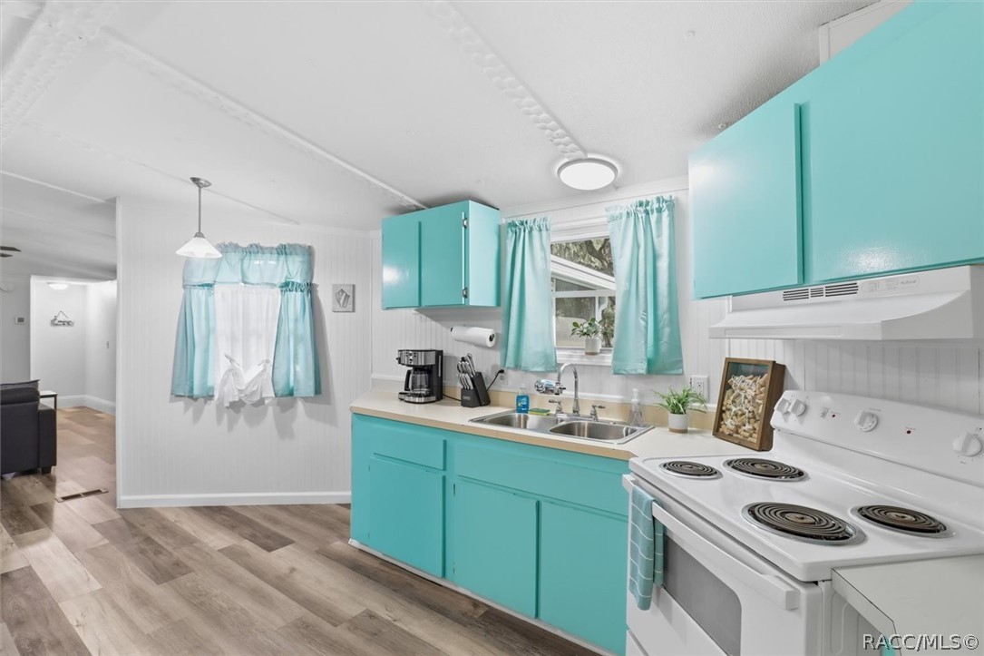 Kitchen with turquoise cabinets, white appliances, and a double-basin sink beneath a curtained window.