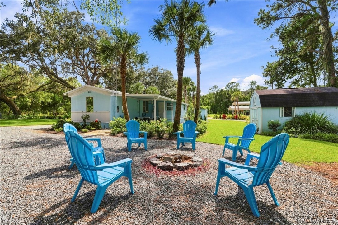 Light blue manufactured home beside a gravel fire pit area with blue Adirondack chairs and palm trees.