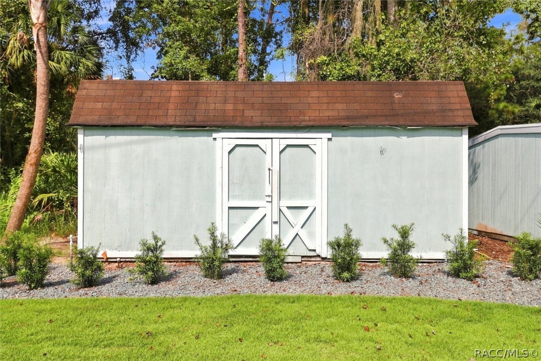 Pale green storage shed with white double doors, brown shingle roof, and shrubs beside a lawn.