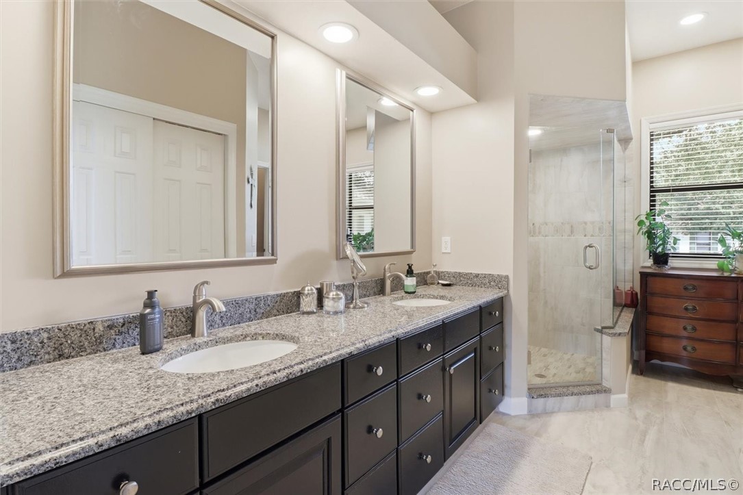 Bathroom with a long double vanity, dark cabinetry, granite counters, and a glass-enclosed shower.