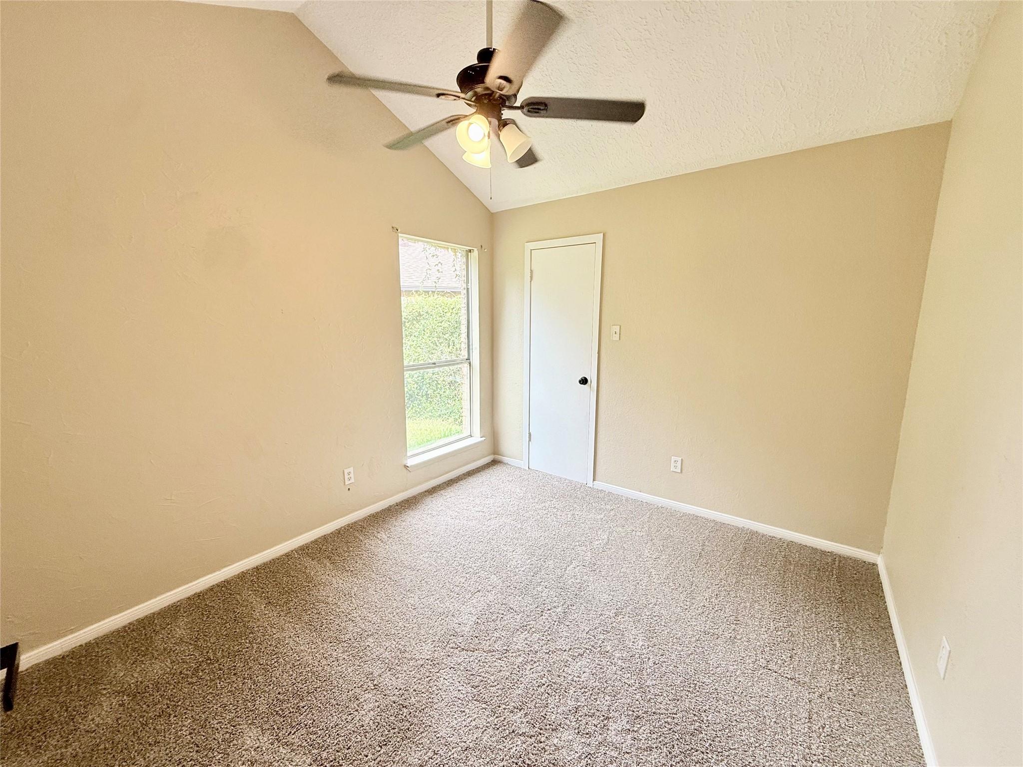 Empty carpeted room with a ceiling fan, sloped ceiling, and window overlooking greenery.