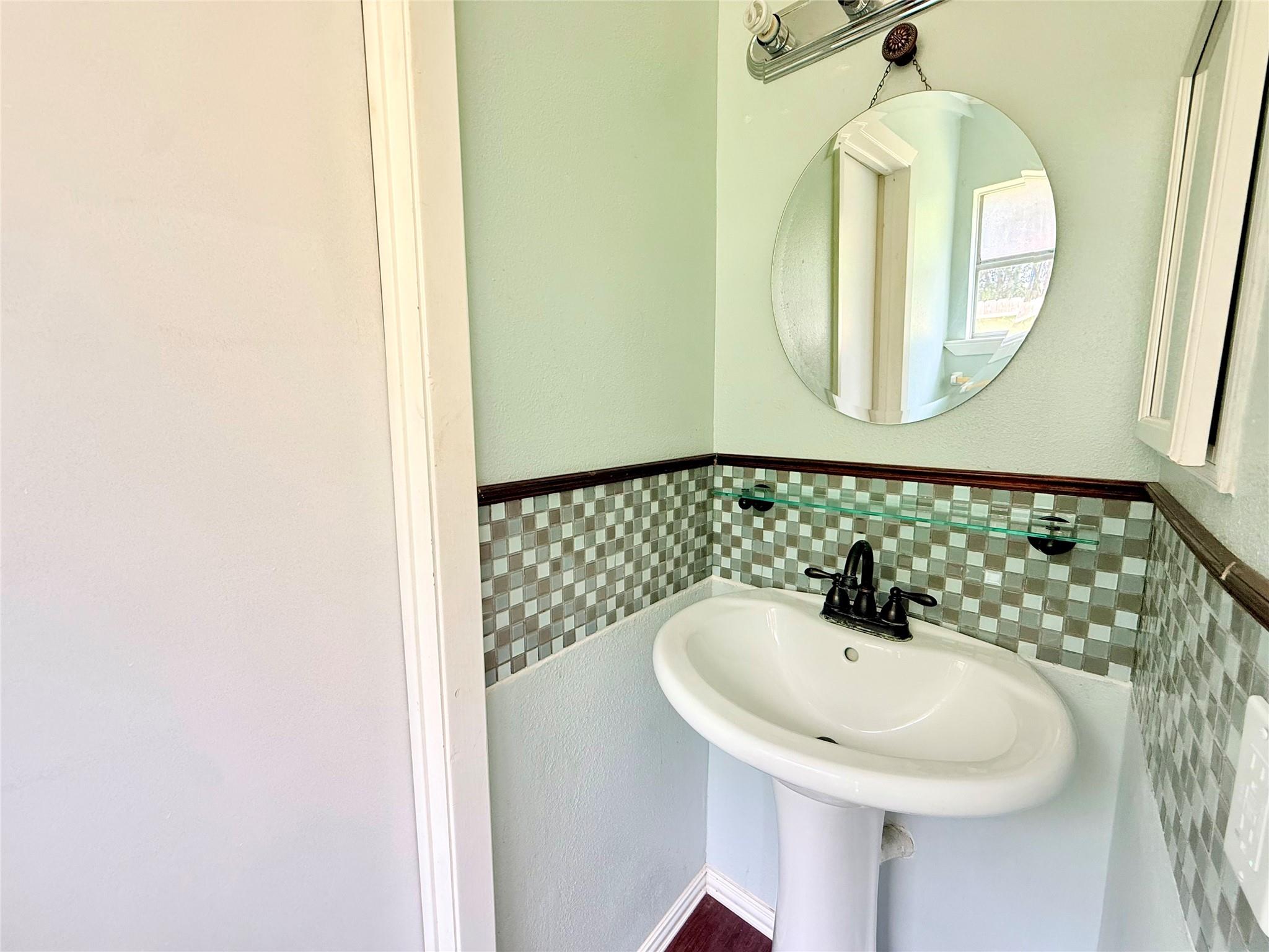 Bathroom pedestal sink with checkerboard tile backsplash, glass shelf, and round mirror.
