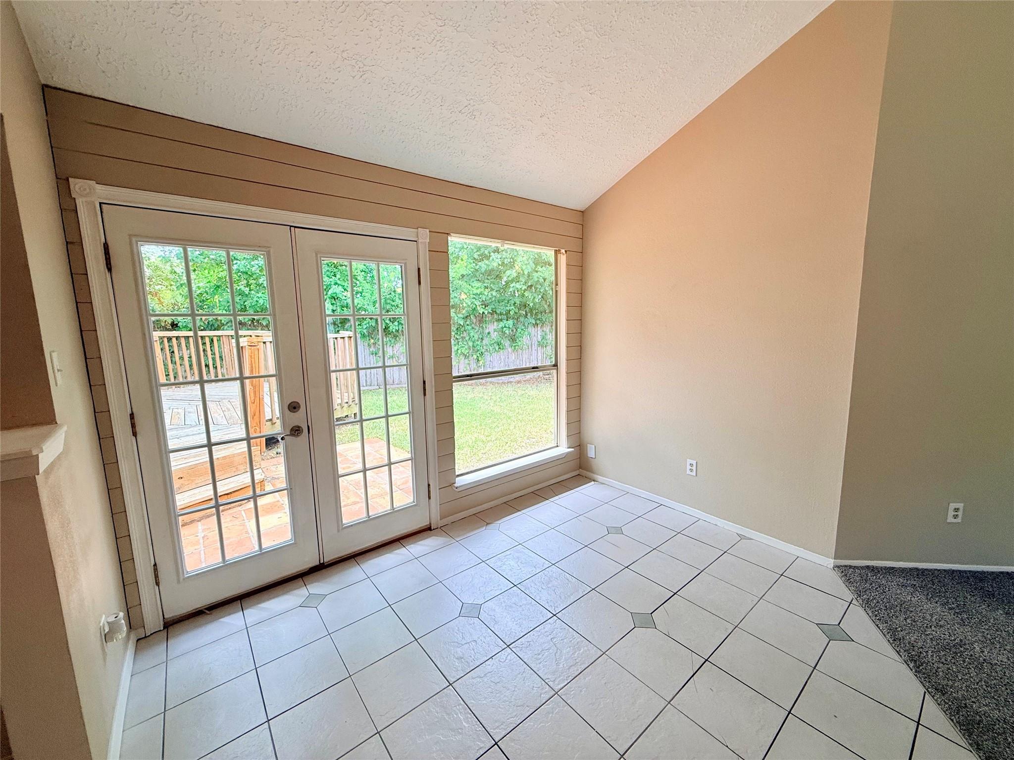 Empty tiled room with French doors opening to a deck and a window overlooking the backyard.