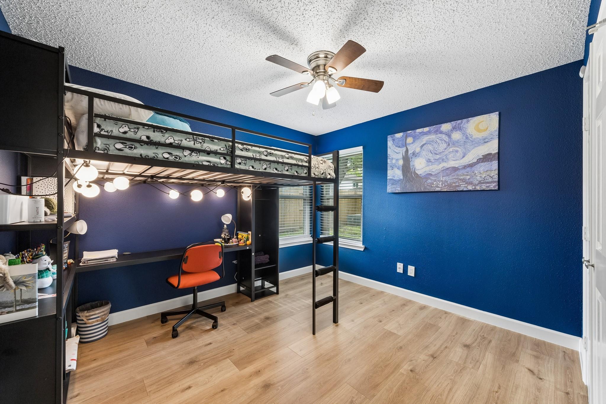 Blue bedroom with a black loft bed and desk, orange chair, and light wood flooring.