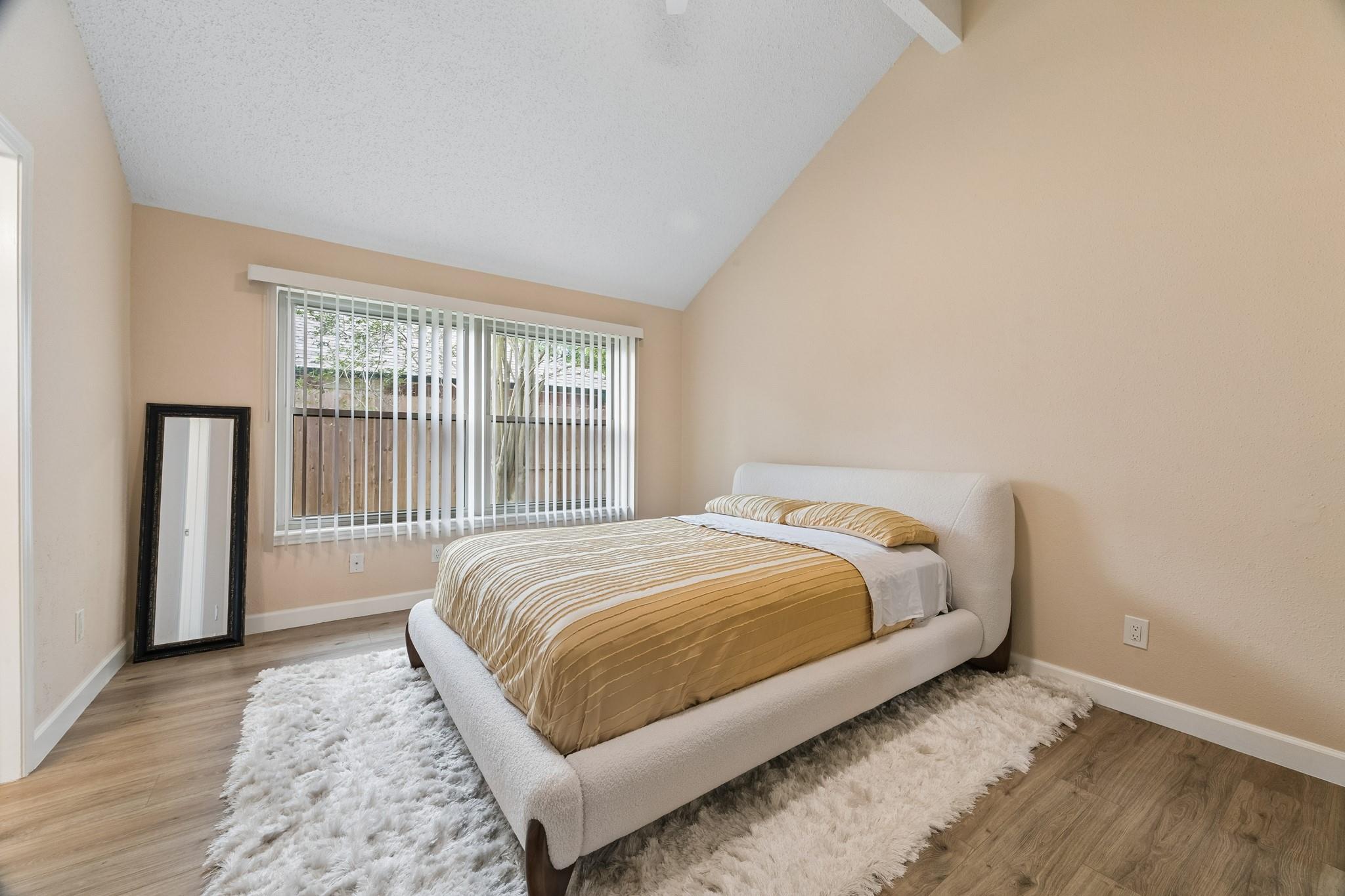 Staged bedroom with a cream bed, gold bedding, sloped ceiling, and large window with vertical blinds.